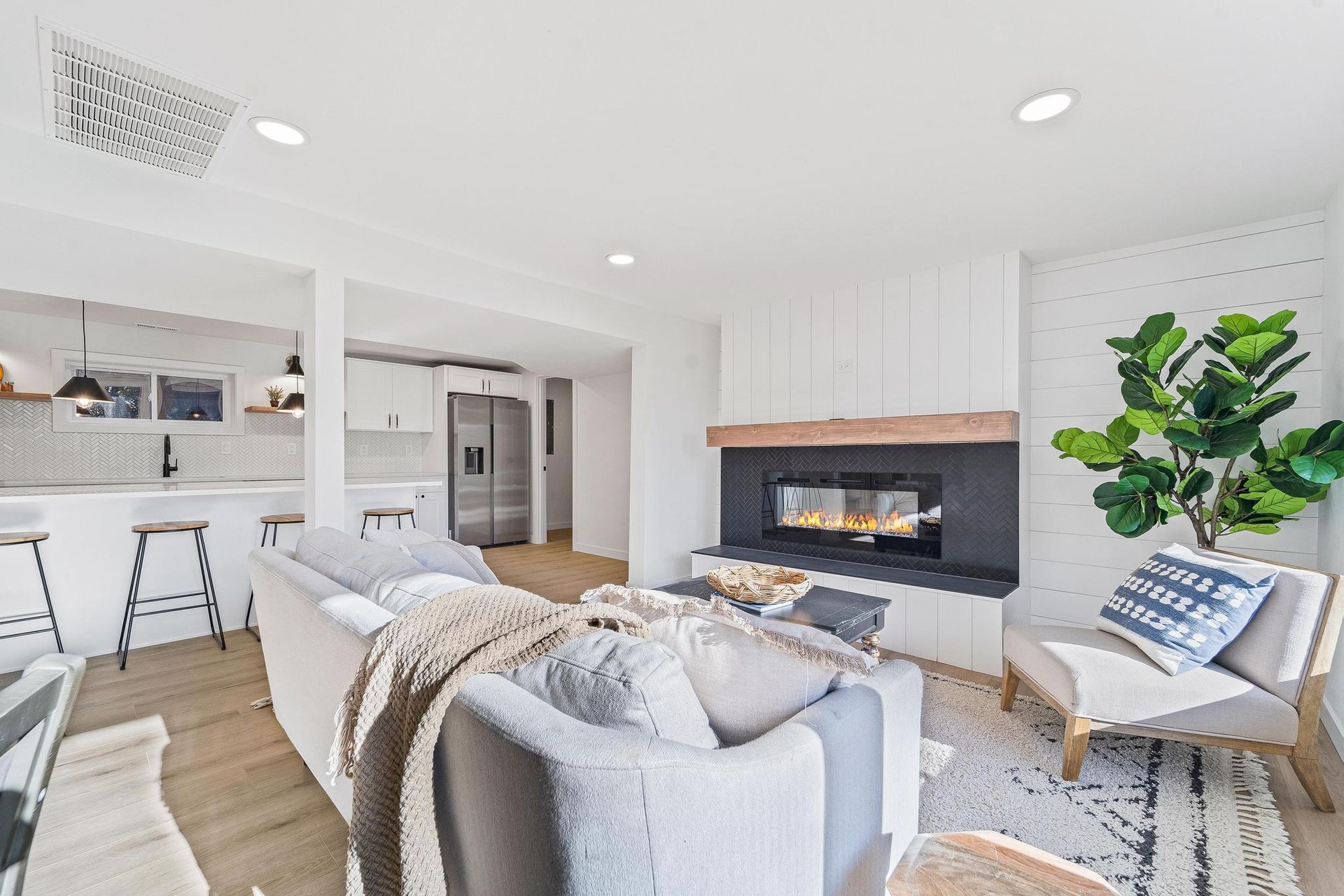 Bright modern living room with fireplace, gray sofa, white chairs, and kitchen opening nearby