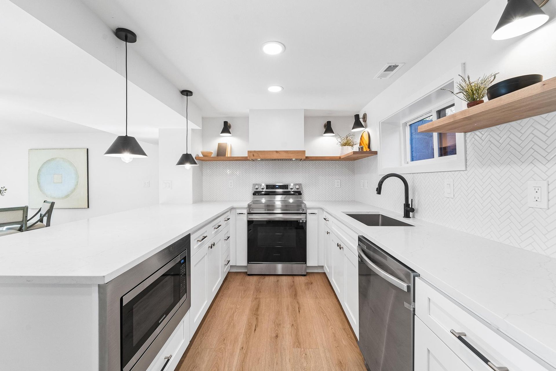 Bright white galley kitchen with black appliances, wood floor, and floating shelves