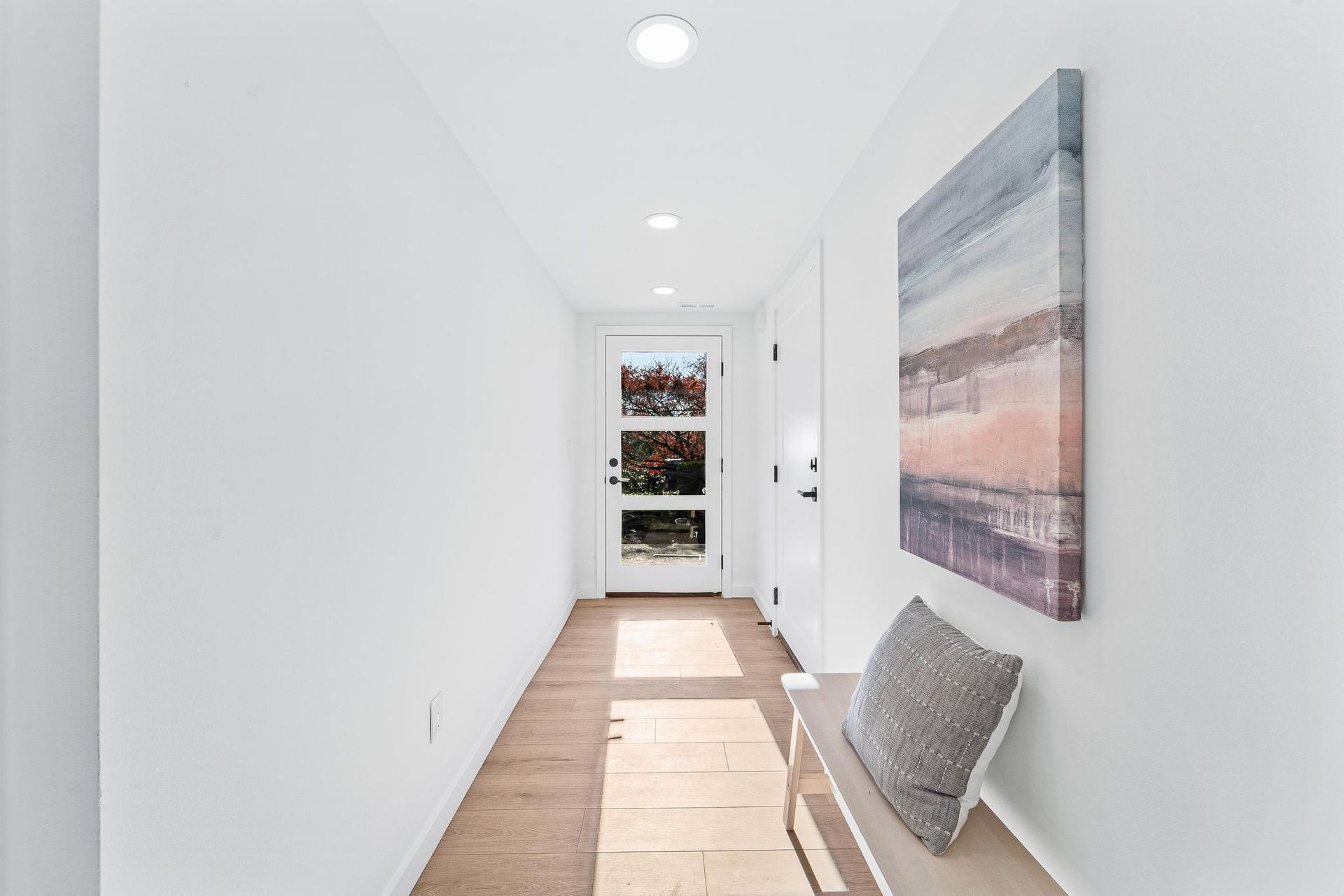 Bright narrow hallway with white walls, recessed lights, a bench, and art leading to a glass door.