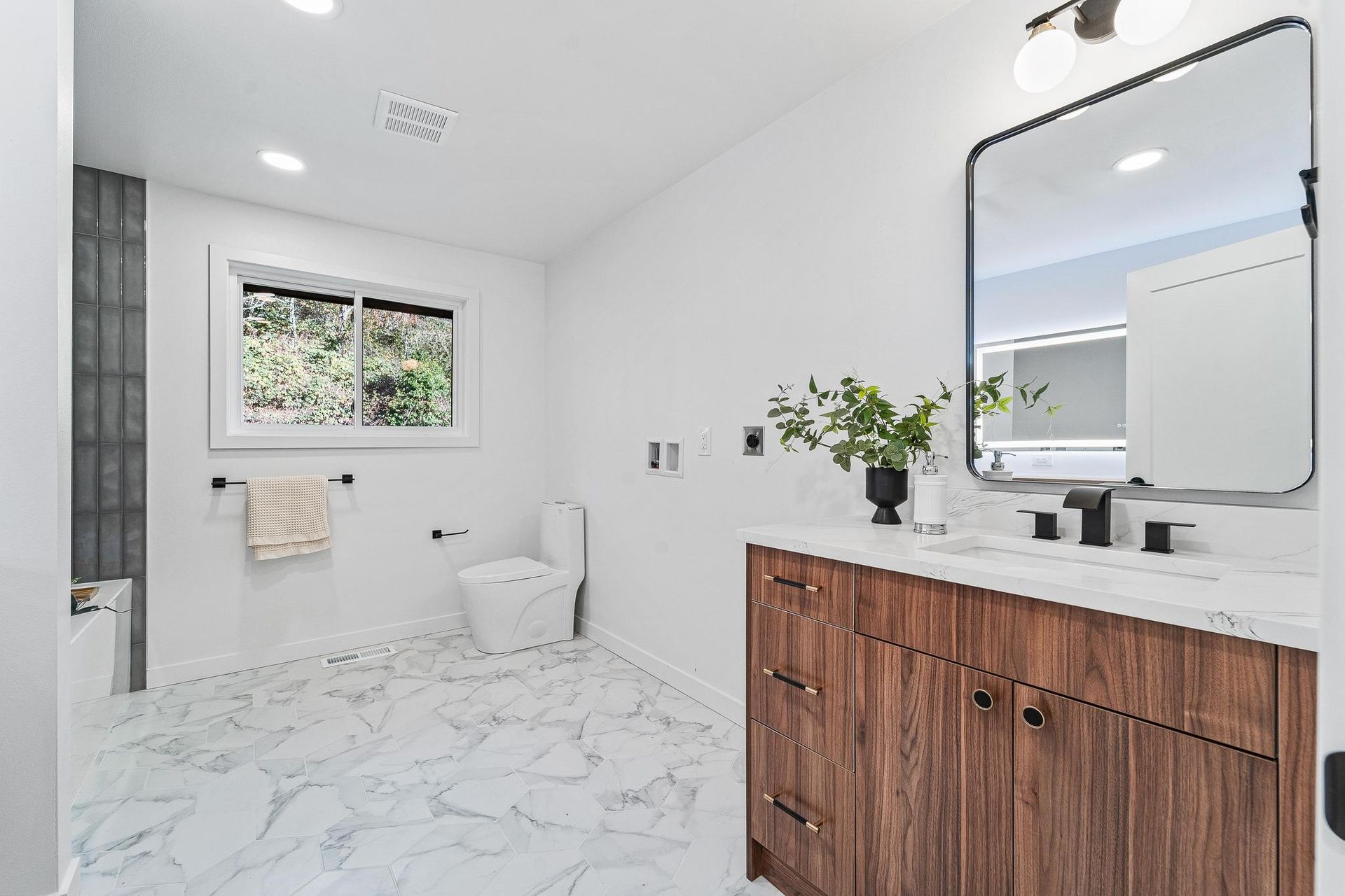 Bright modern bathroom with white walls, marble tile, wood vanity, mirror, toilet, and window