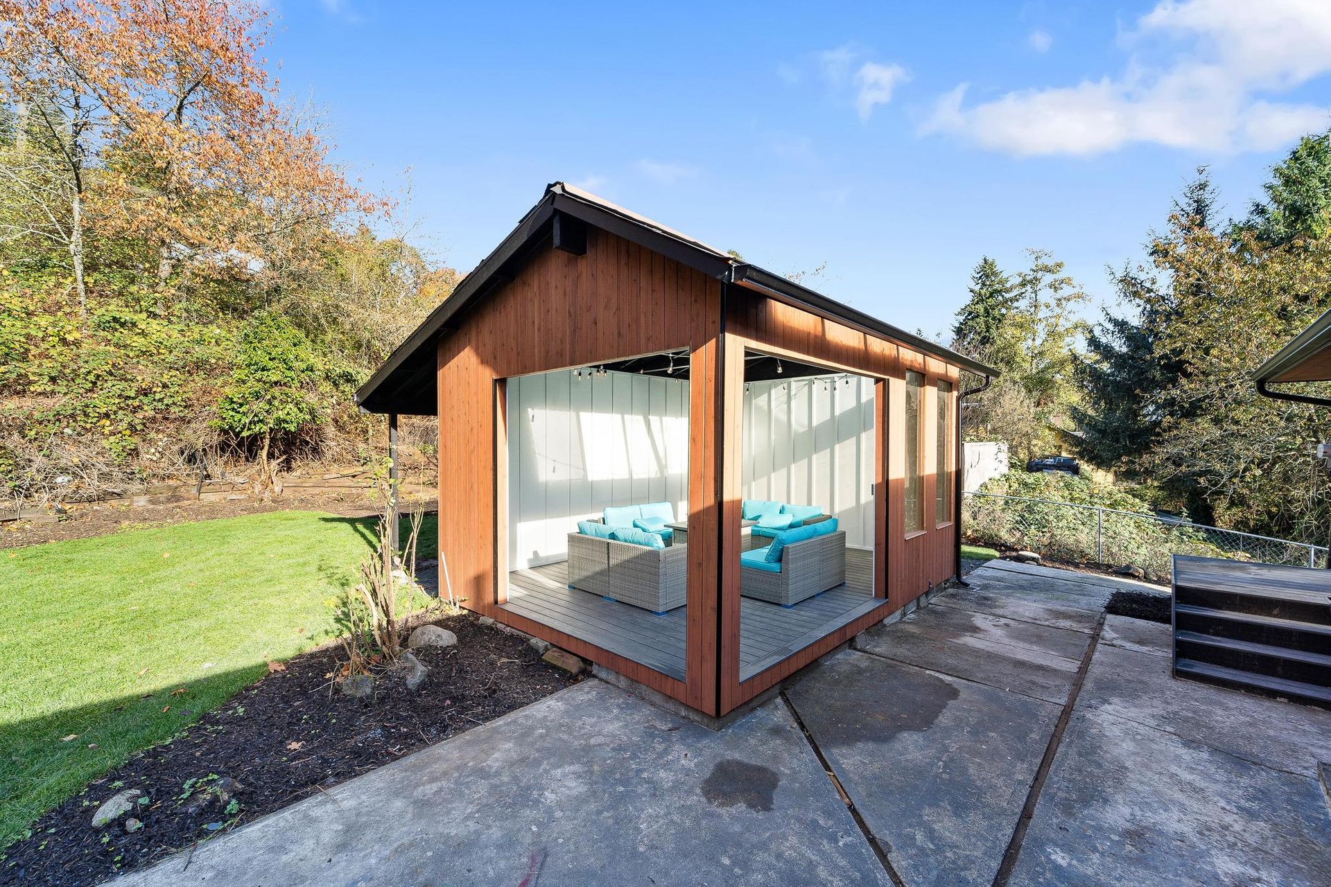 Glass-walled backyard sauna with wooden frame, stone patio, and steps in a sunny garden