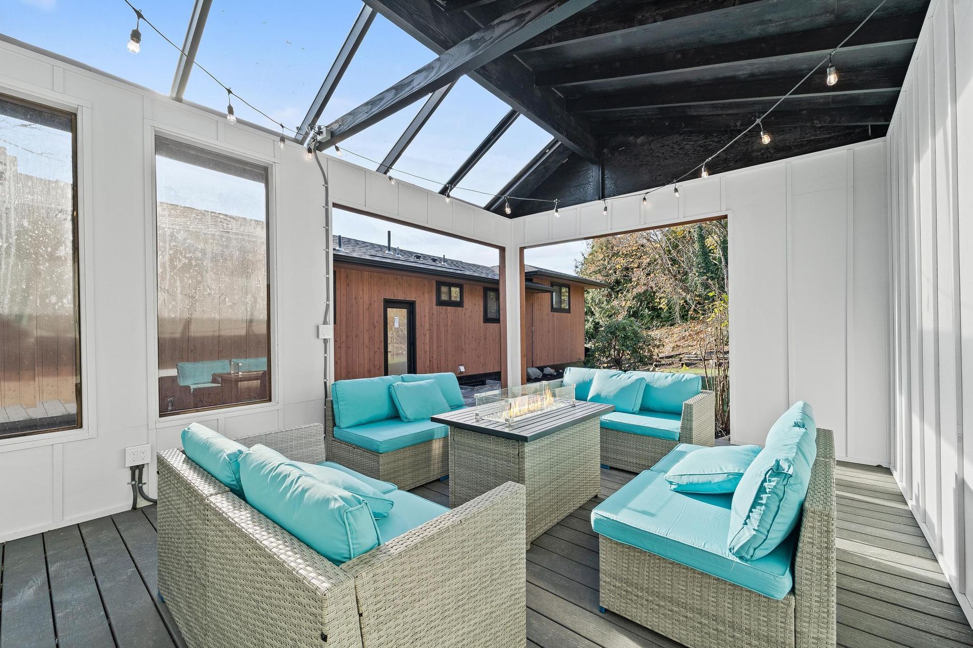 Covered patio with turquoise-cushioned wicker seating and a glass roof, overlooking a backyard.