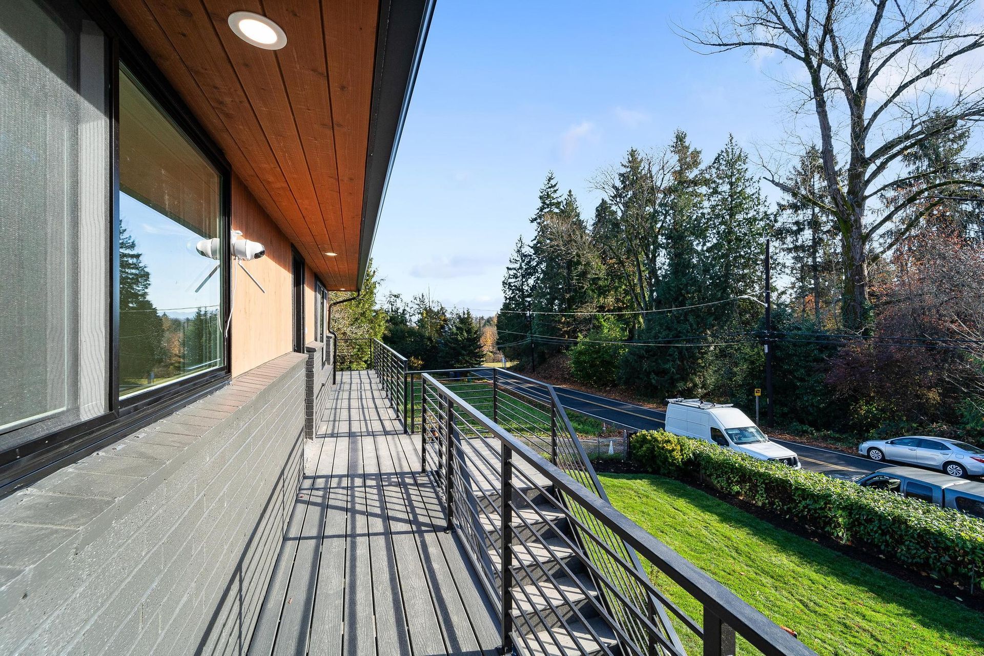 Balcony walkway beside a building, with metal railing, green lawn, trees, and parked cars below.