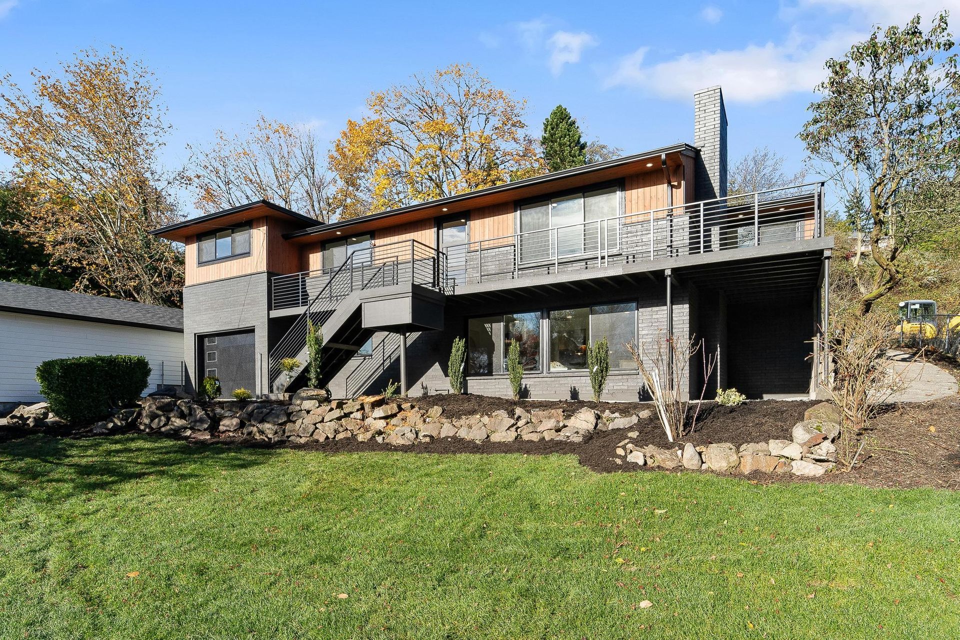 Two-story house with gray deck and stairs on a grassy hill under autumn trees