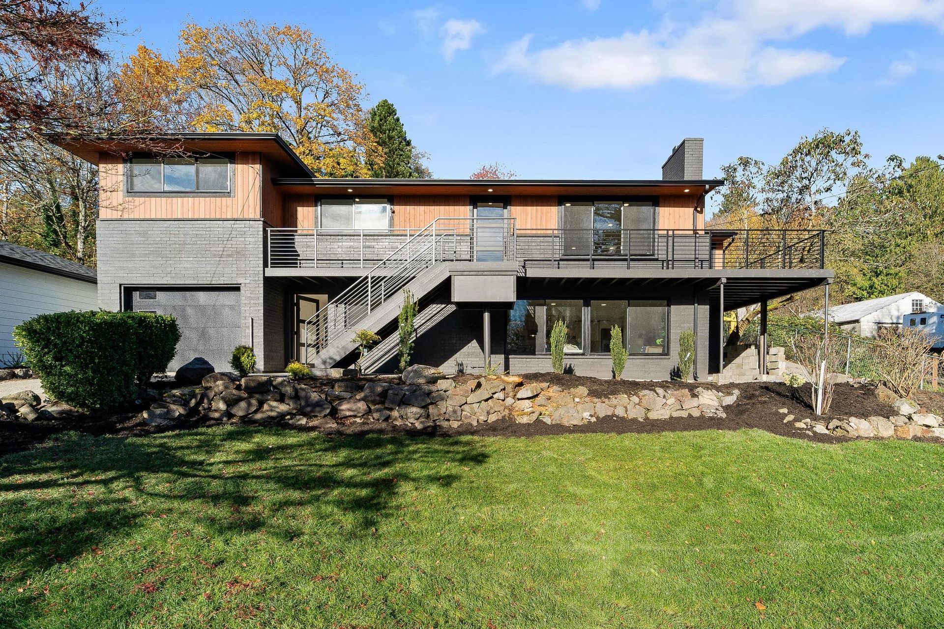 Backyard view of a two-story modern house with stone lower level, deck, stairs, and green lawn