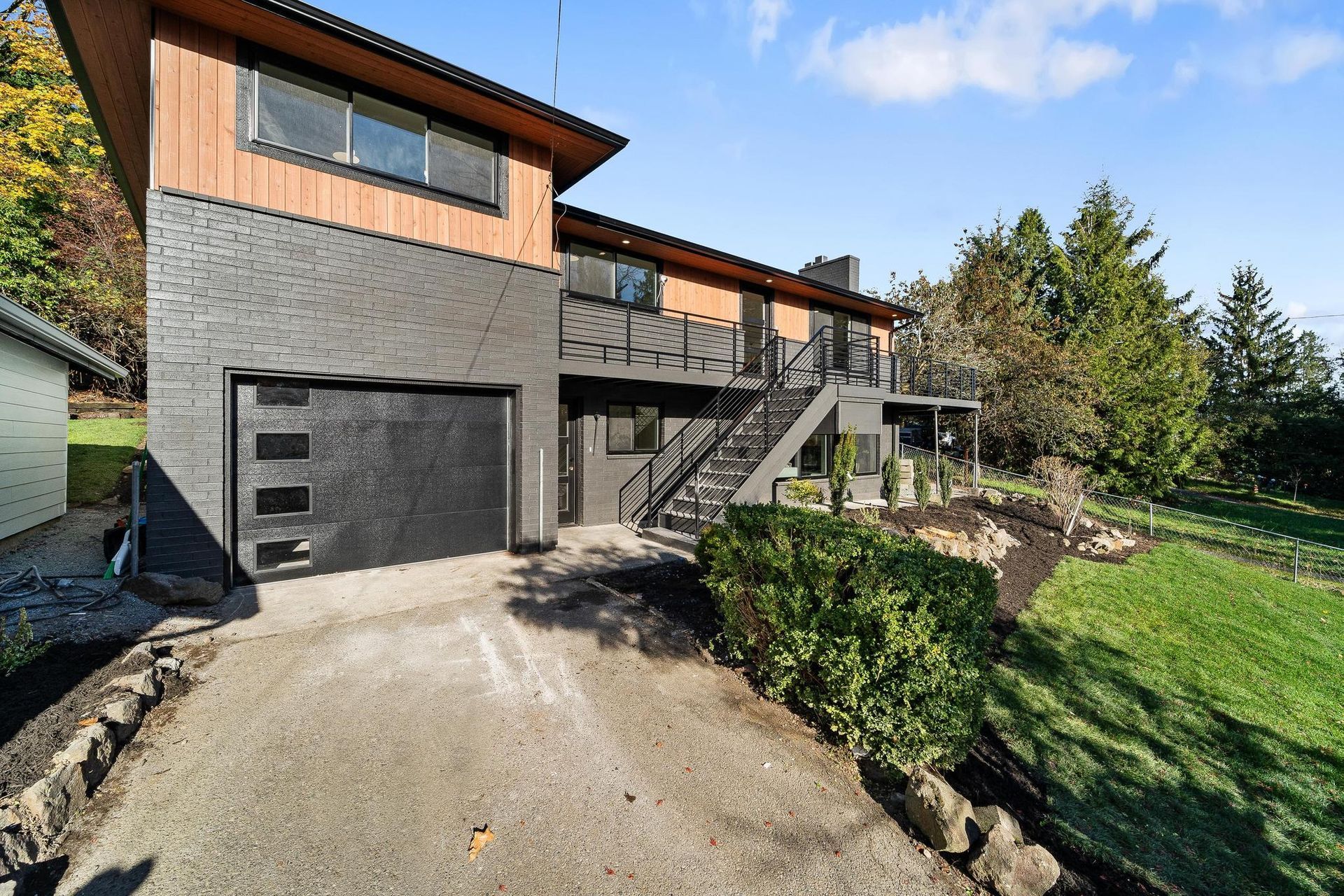 Modern two-story house with black garage, exterior stairs, and sloped driveway surrounded by trees