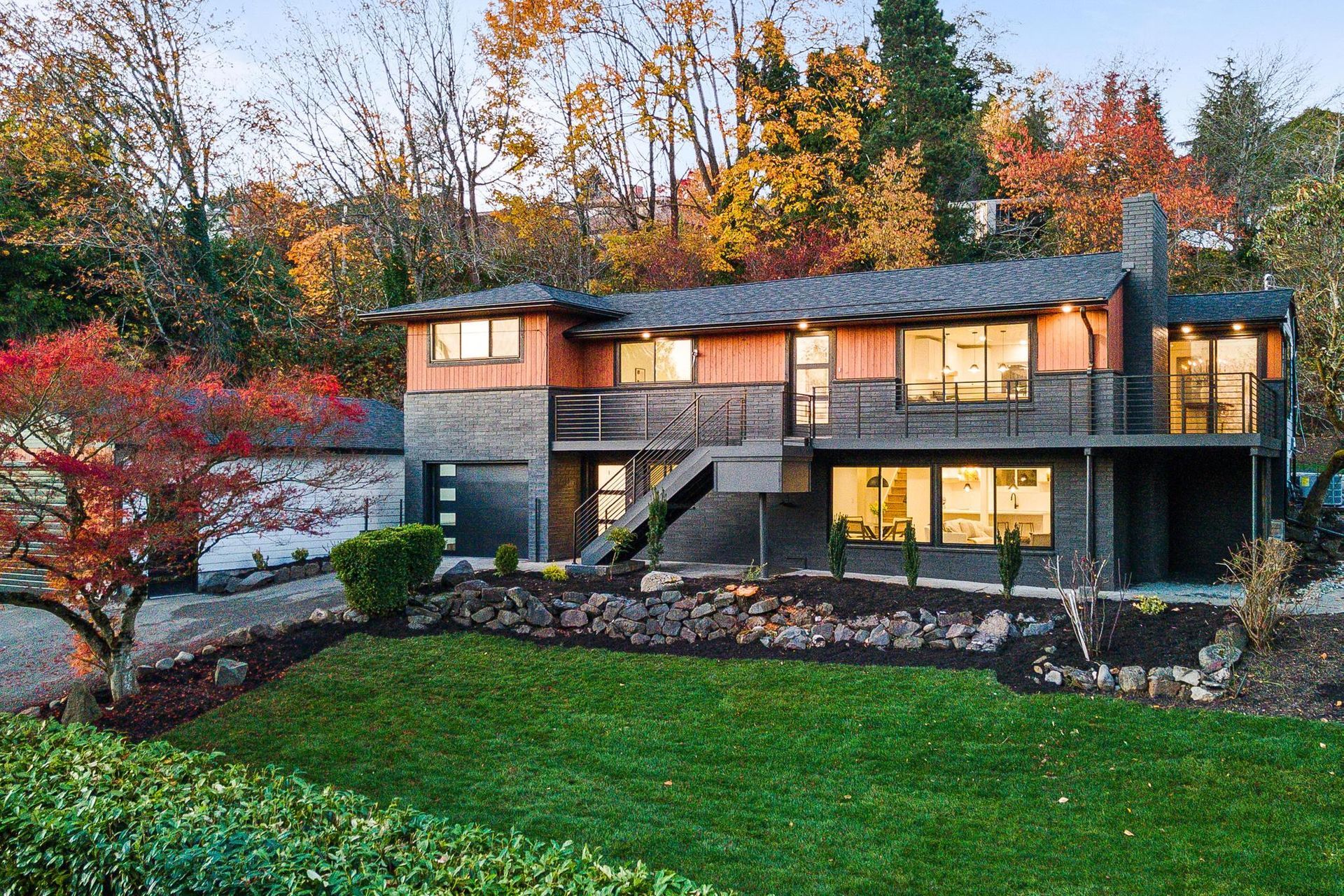 Modern two-story house with lit windows, stone accents, and autumn trees in front yard