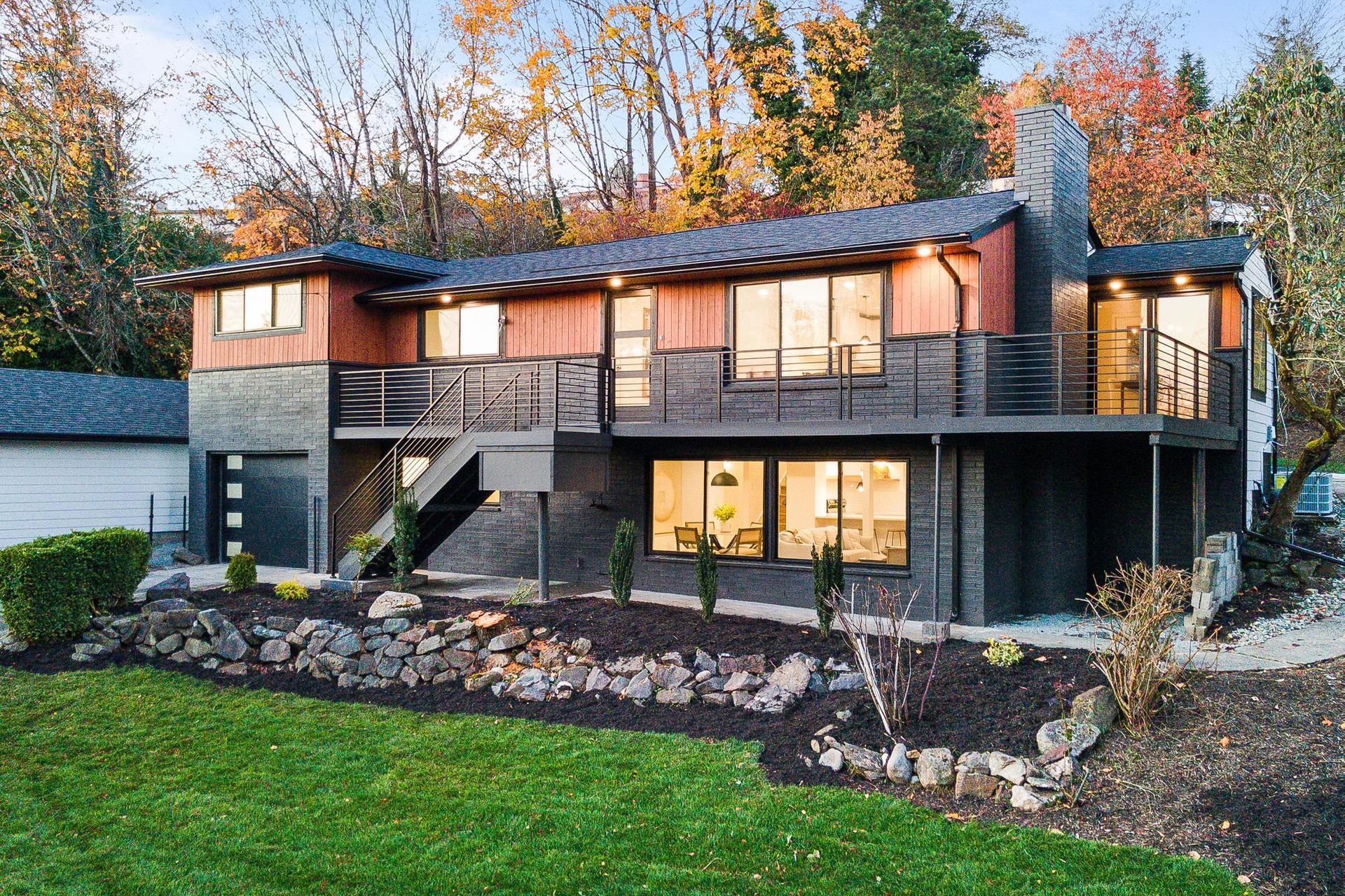 Modern two-story house with dark siding, large windows, and a landscaped front yard at dusk