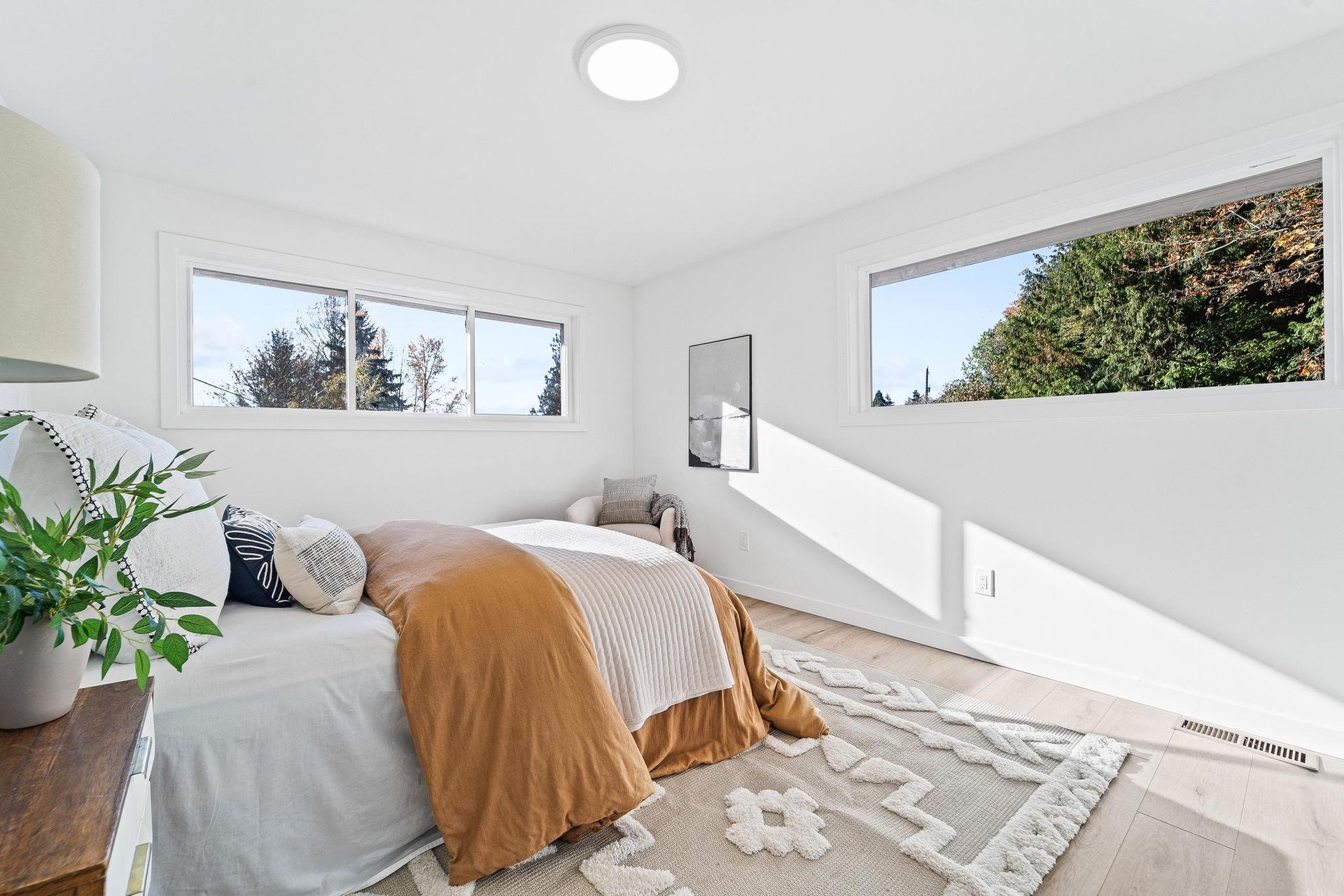 Bright bedroom with large windows, a bed with orange throw, and a potted plant by the window.