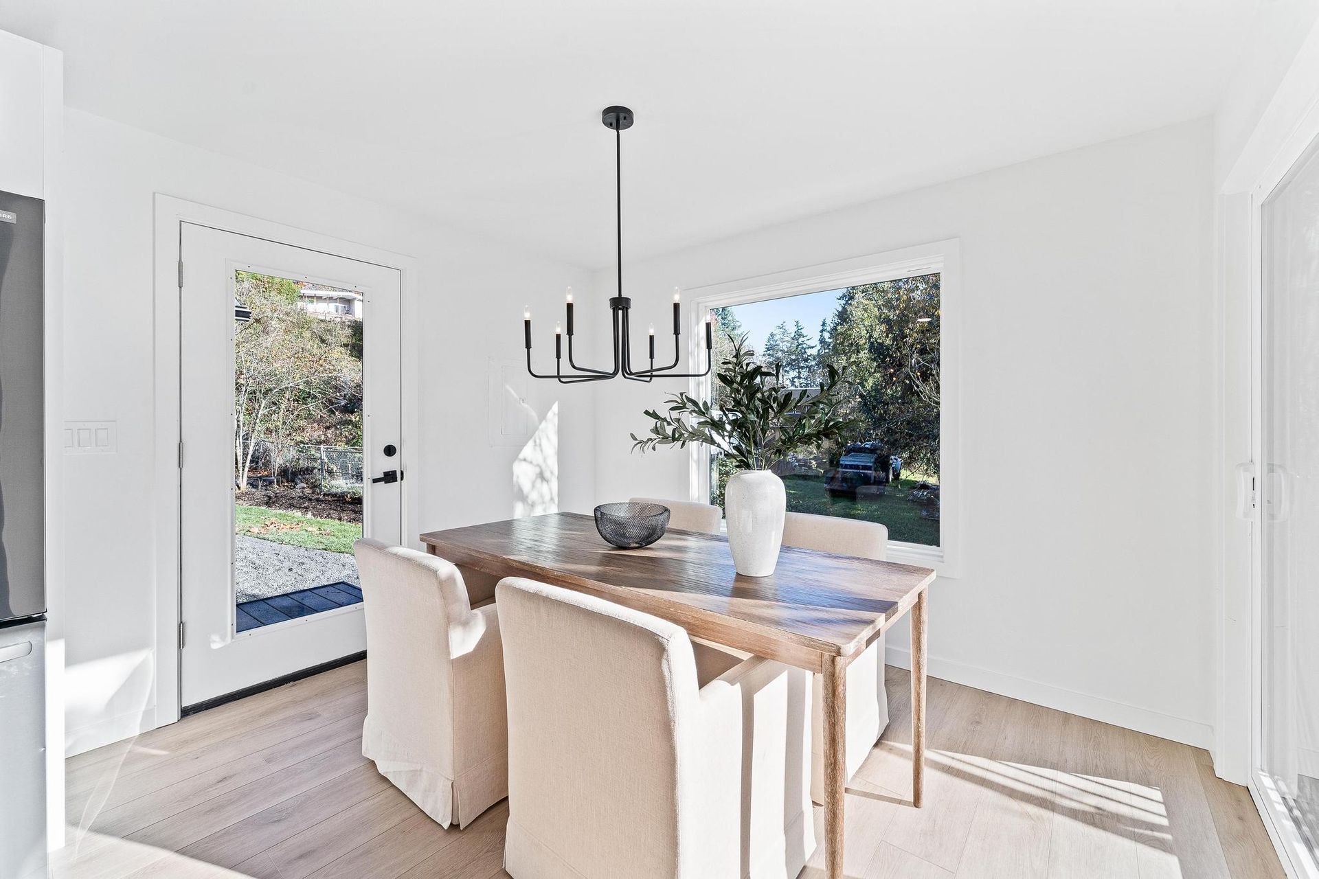 Bright dining room with a wooden table, cream chairs, black chandelier, and large windows