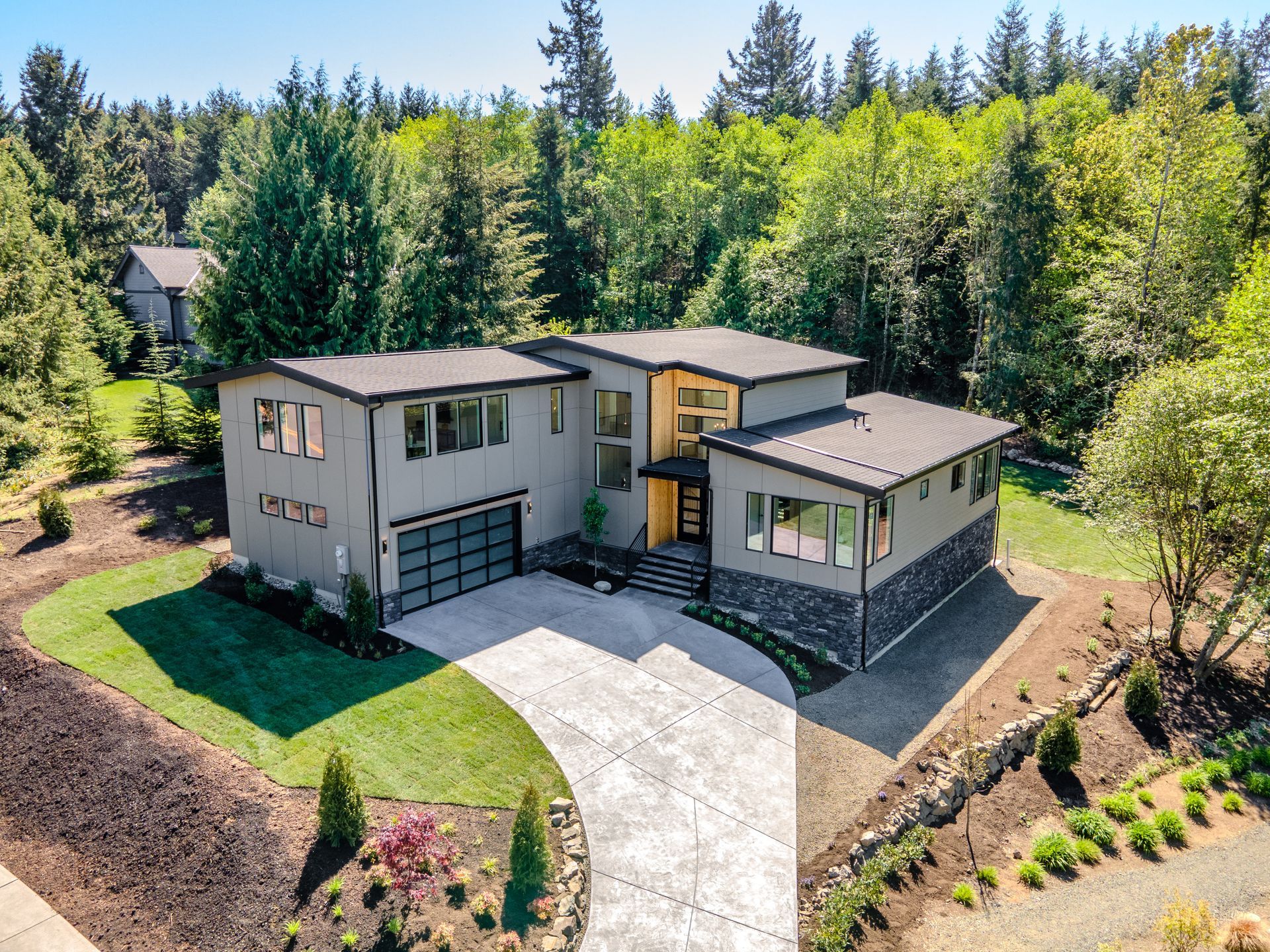 Modern two-story house with light siding, dark roof, and curved driveway in a wooded setting