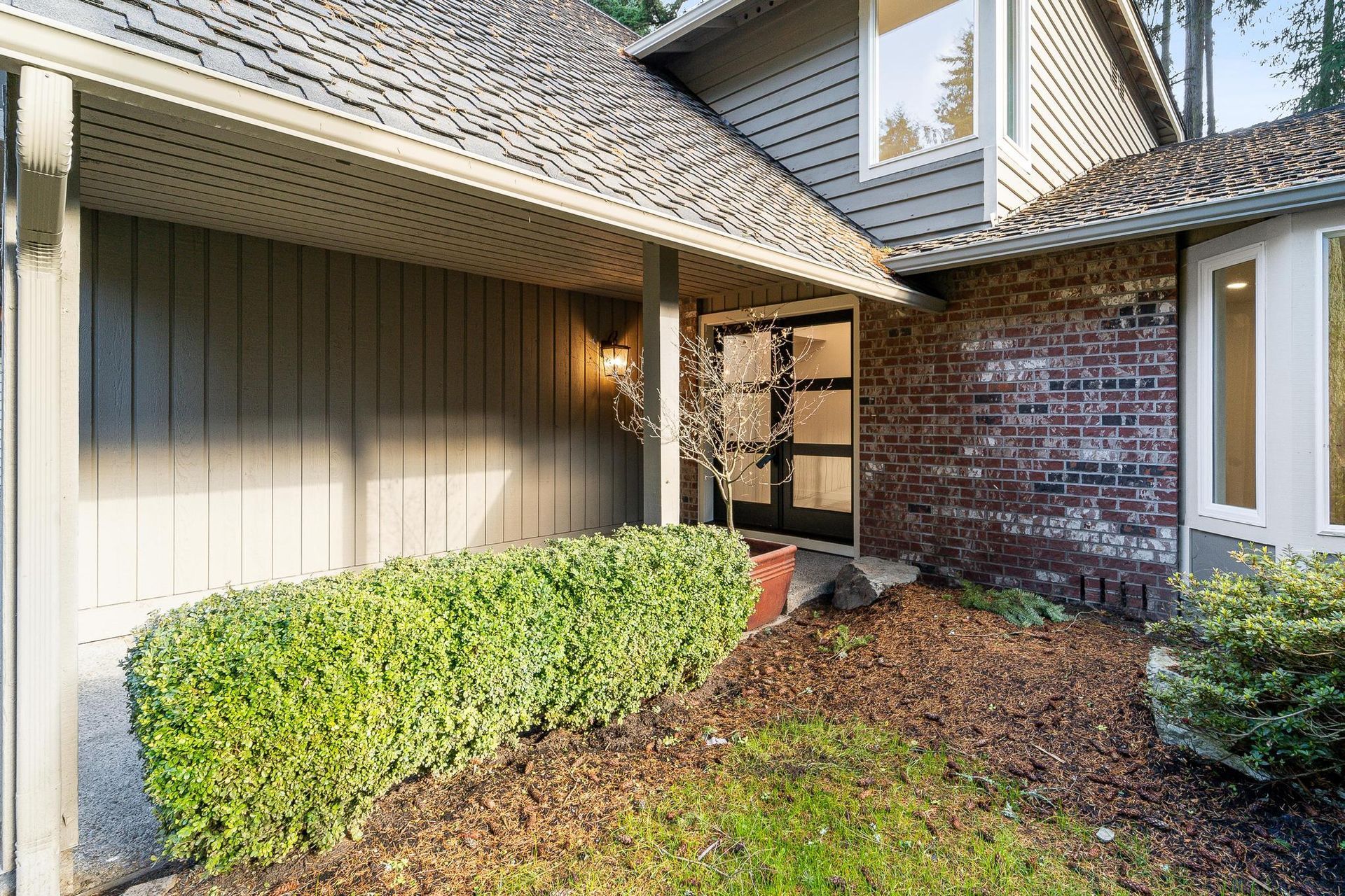Front entry patio with hedges, brick wall, and house siding under a shingled roof
