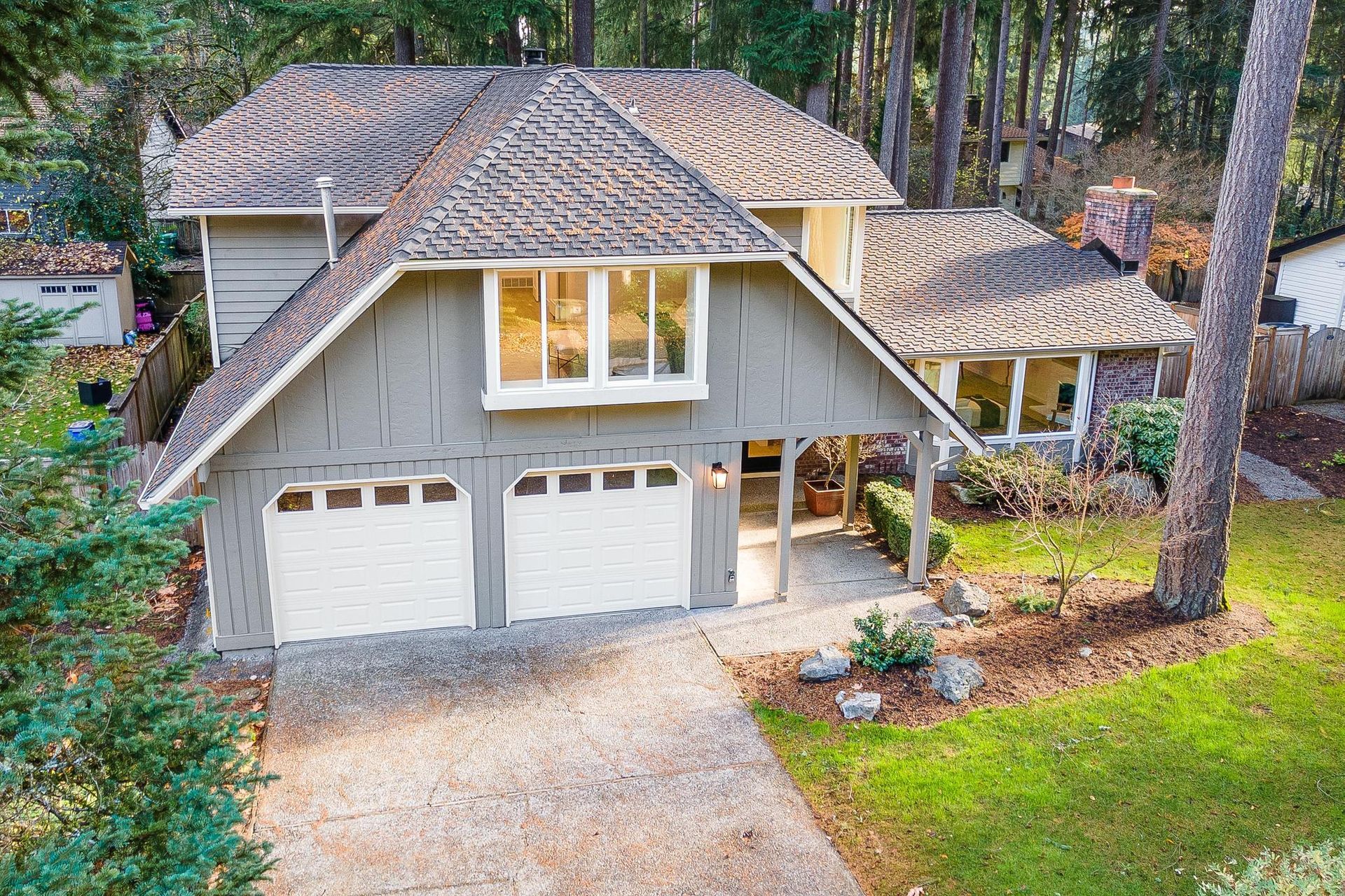 Aerial view of a gray house with a two-car garage, gravel driveway, and wooded yard