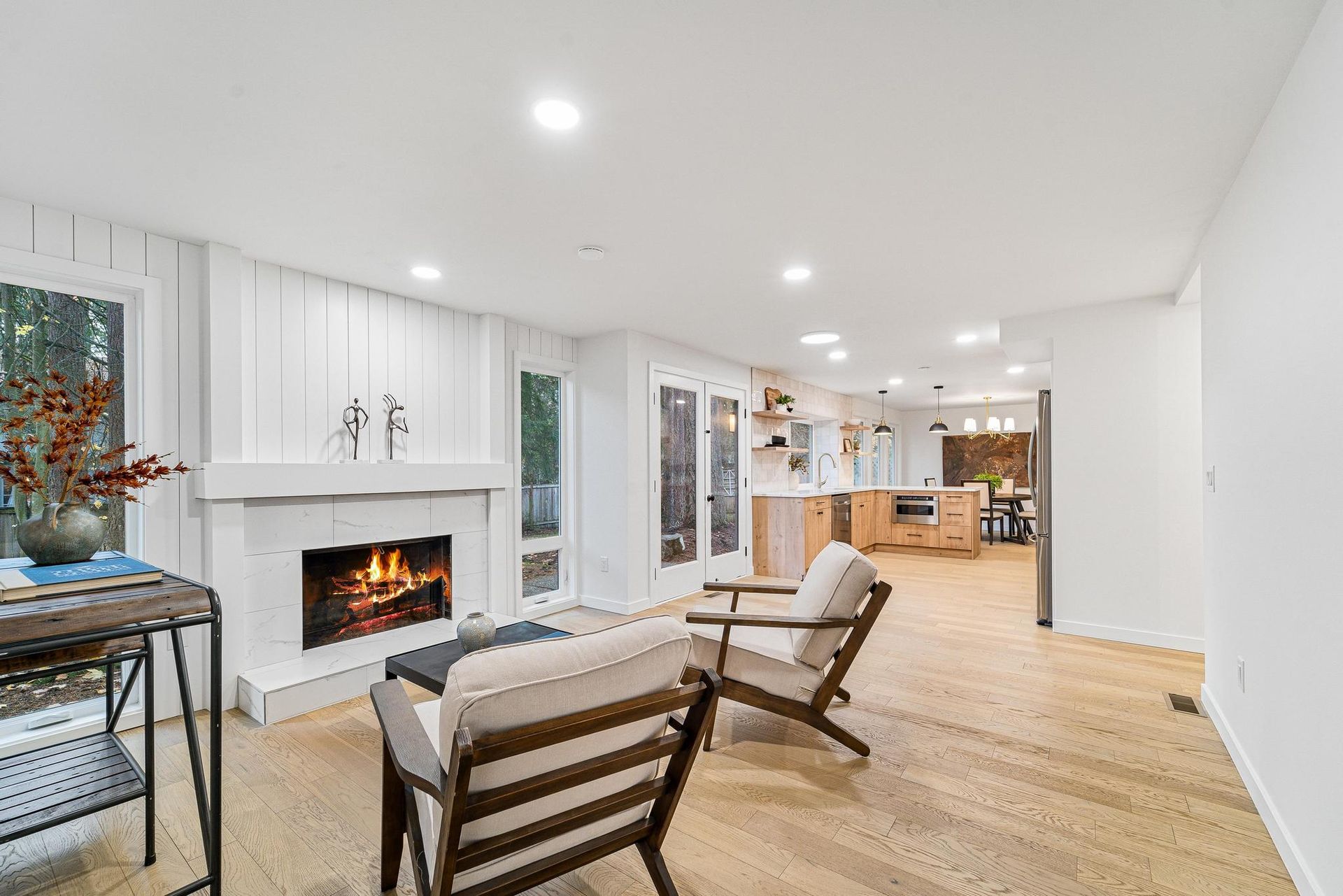 Bright open living room with fireplace, white walls, wood floors, and a kitchen in the background