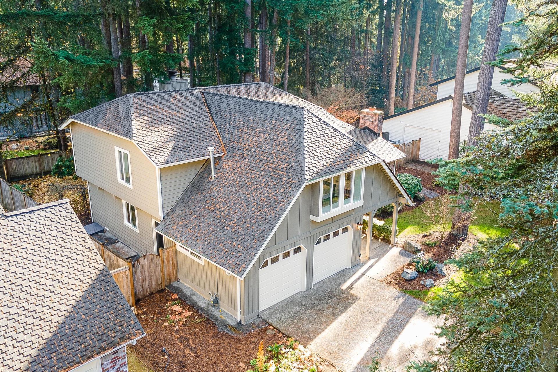 Aerial view of a beige house with gray roof and garage, surrounded by tall trees and a driveway.