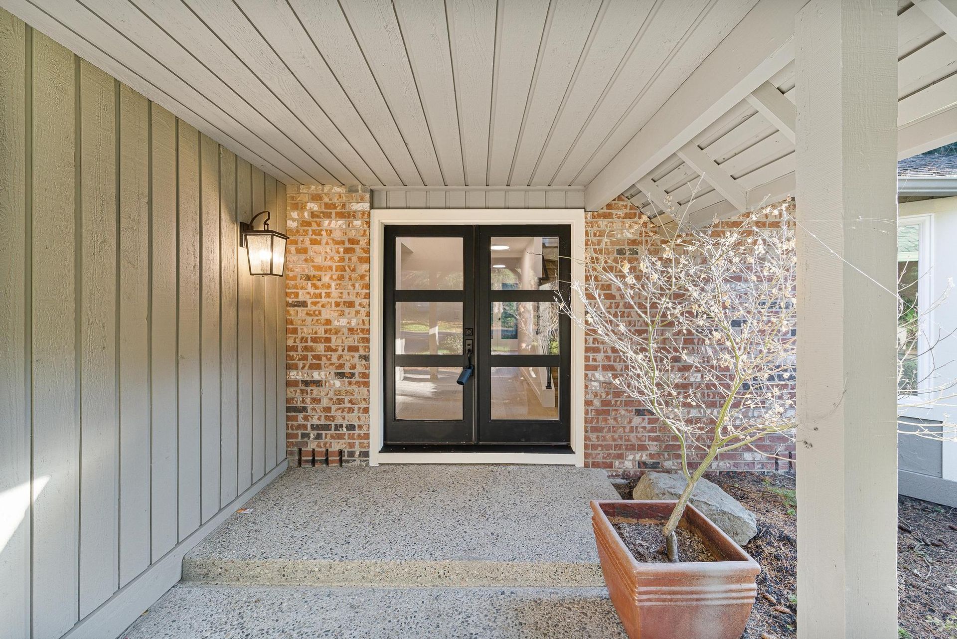 Covered front entry with black double doors, gray siding, brick wall, and a potted plant on the porch