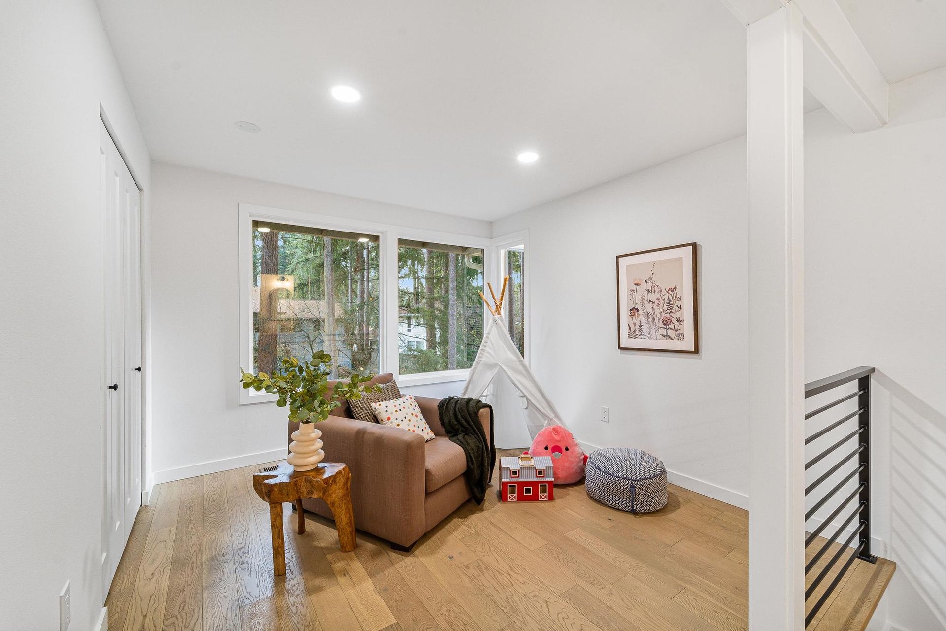 Bright living room with brown armchair, wood floor, window view, and a staircase railing.