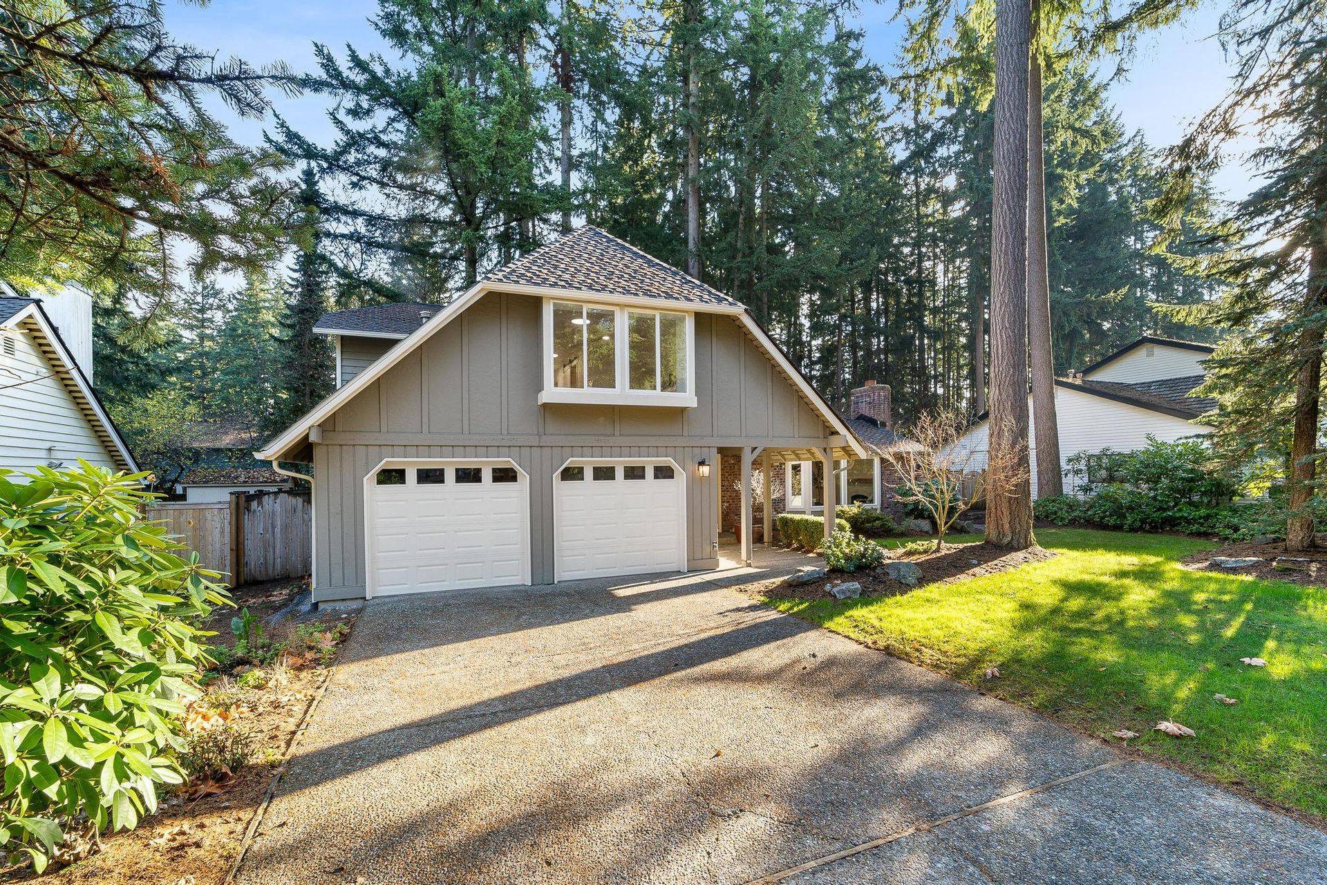 Front view of a gray two-car garage home with gravel driveway and tall trees in the yard