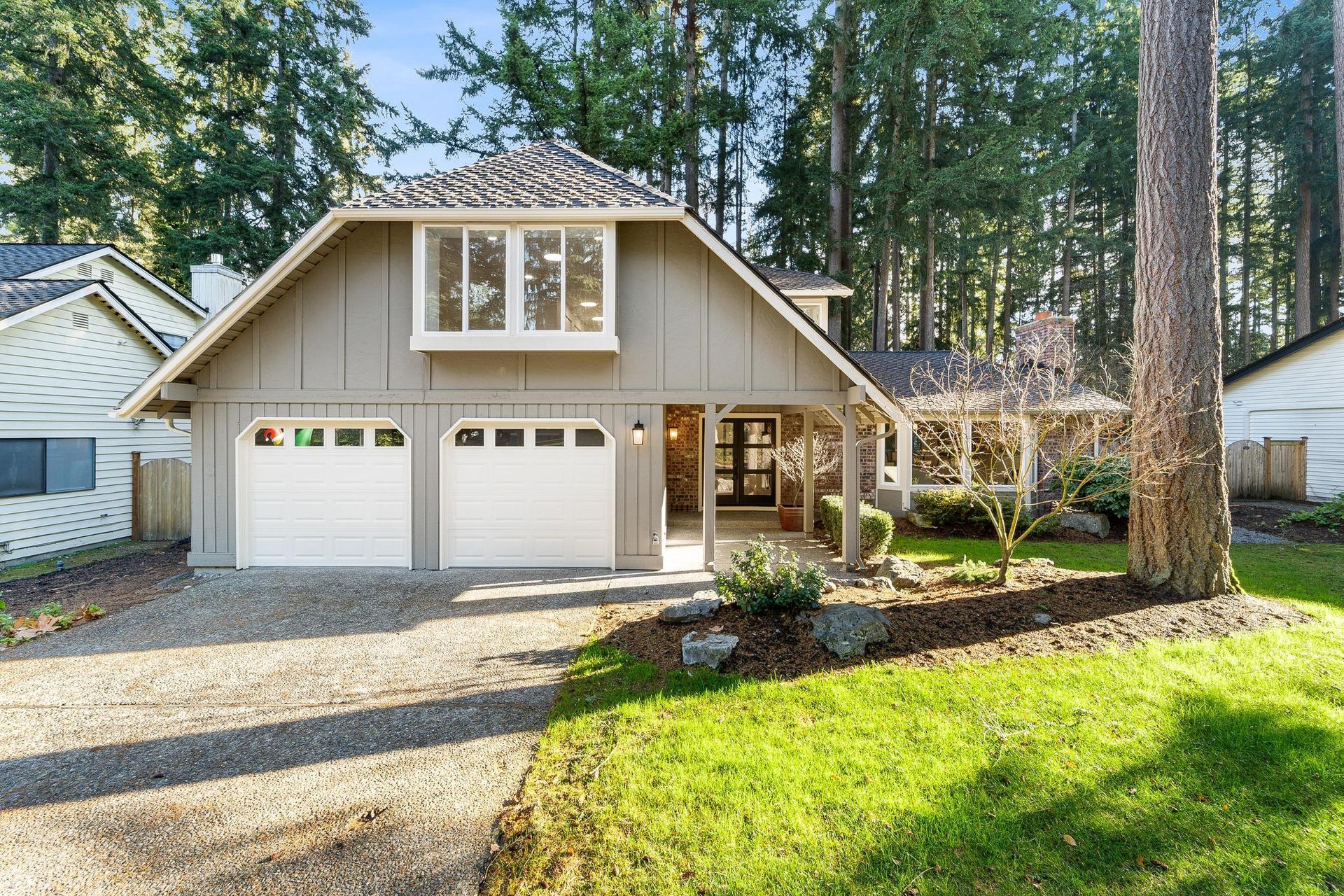 Two-story tan house with double garage, front porch, and tall trees in a sunny yard