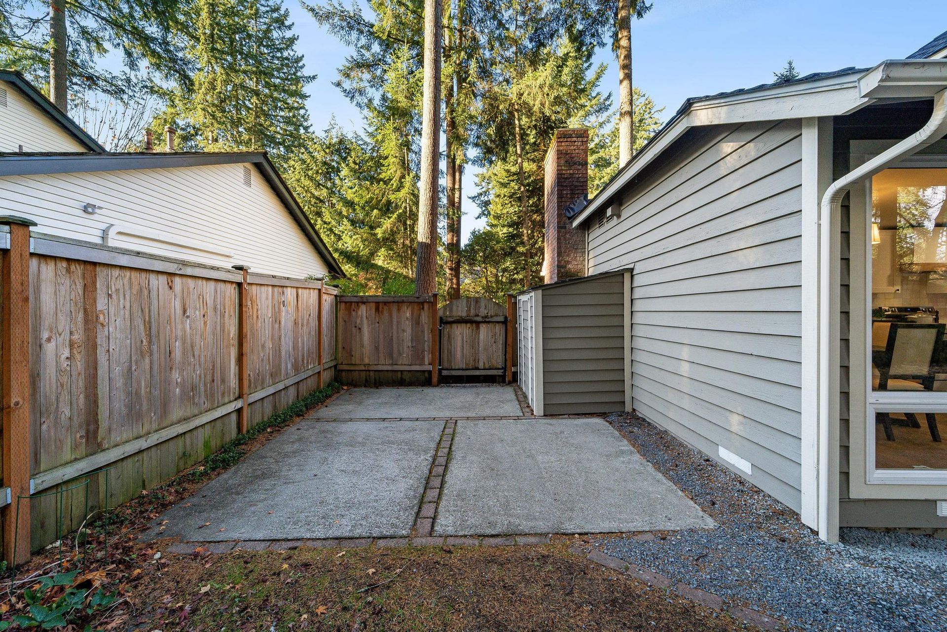Gravel backyard patio beside a gray house and wooden fence, with tall trees in the background