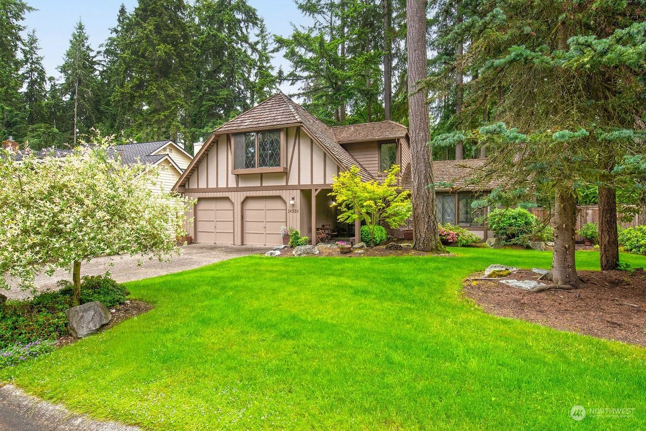 Suburban house with tan siding, two-car garage, and a lush green lawn surrounded by trees.
