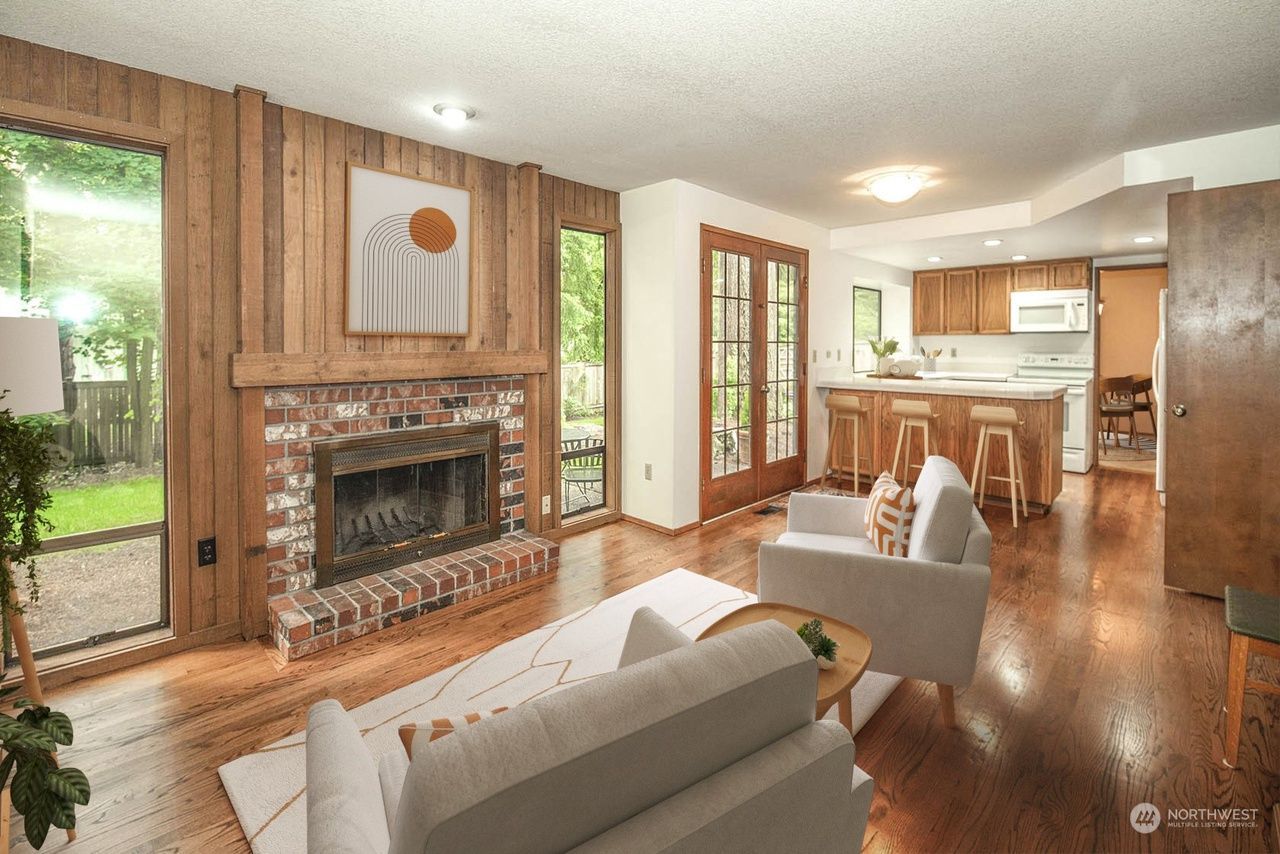 Bright living room with brick fireplace, wood paneling, white chairs, and open kitchen beyond.