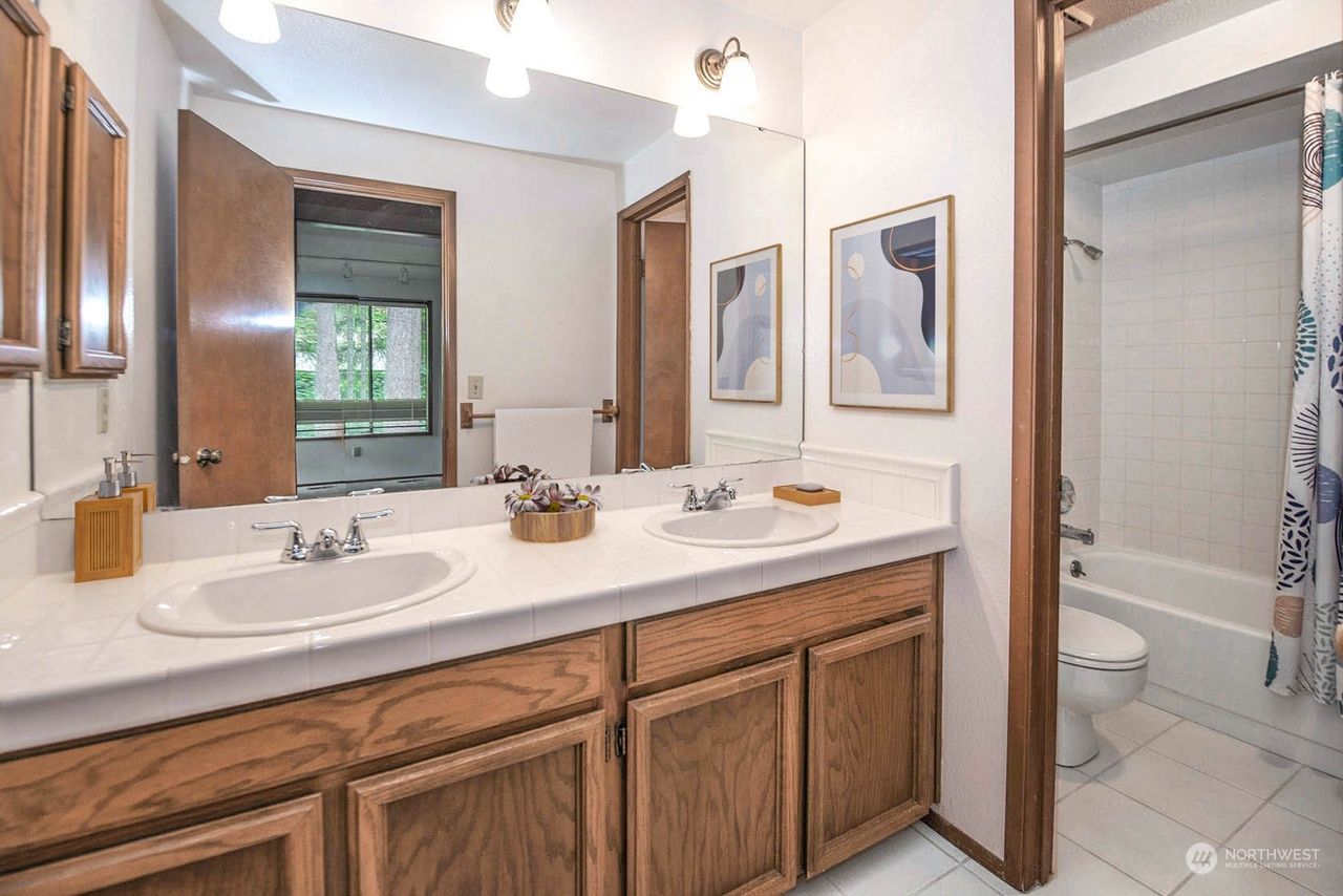 Bathroom vanity with double sinks, wood cabinets, mirror, and a glimpse of a tiled shower and toilet.