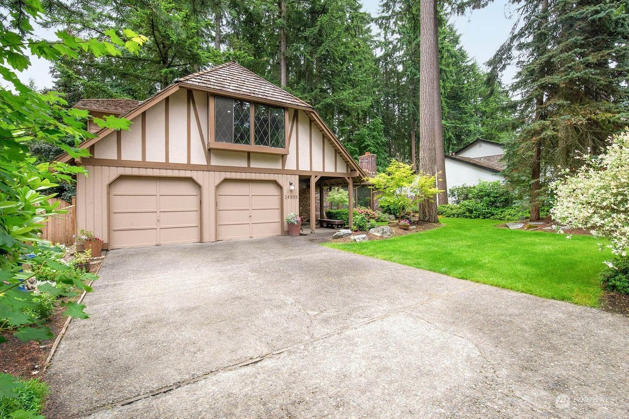 Suburban house with two-car garage, gravel driveway, and lush green trees and lawn