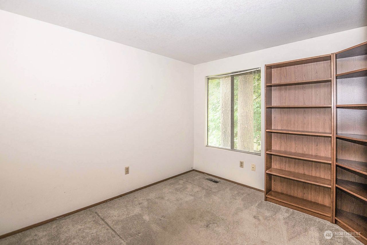 Empty beige bedroom with carpet, window, and tall wooden bookshelf.