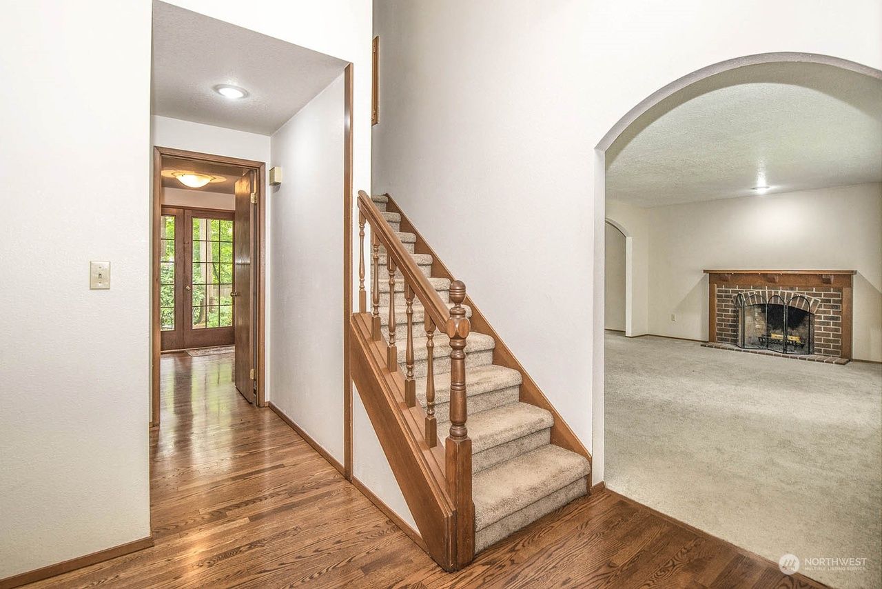 Bright hallway with hardwood floors, staircase, and arched opening to a living room with fireplace.