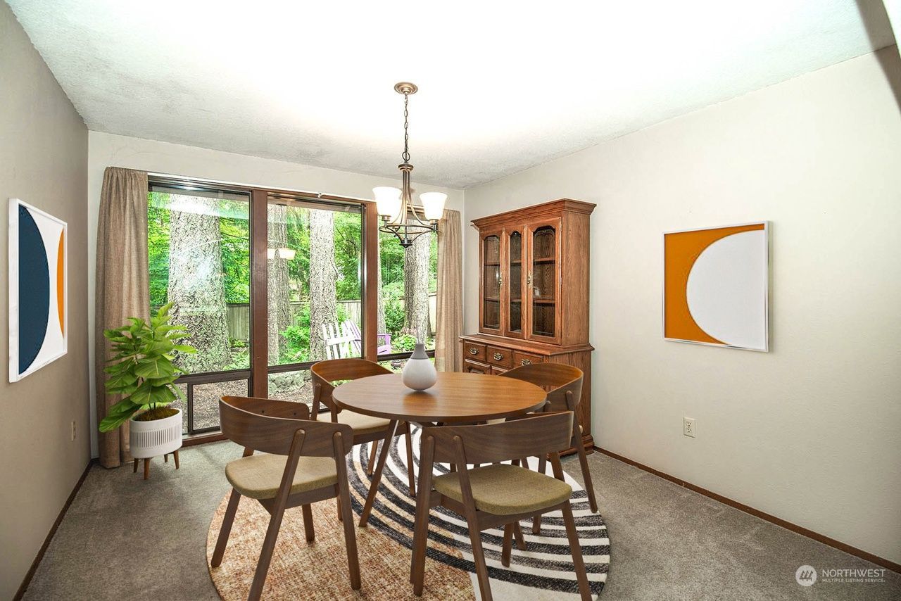 Dining room with round wooden table, chairs, china cabinet, and large windows with greenery outside.