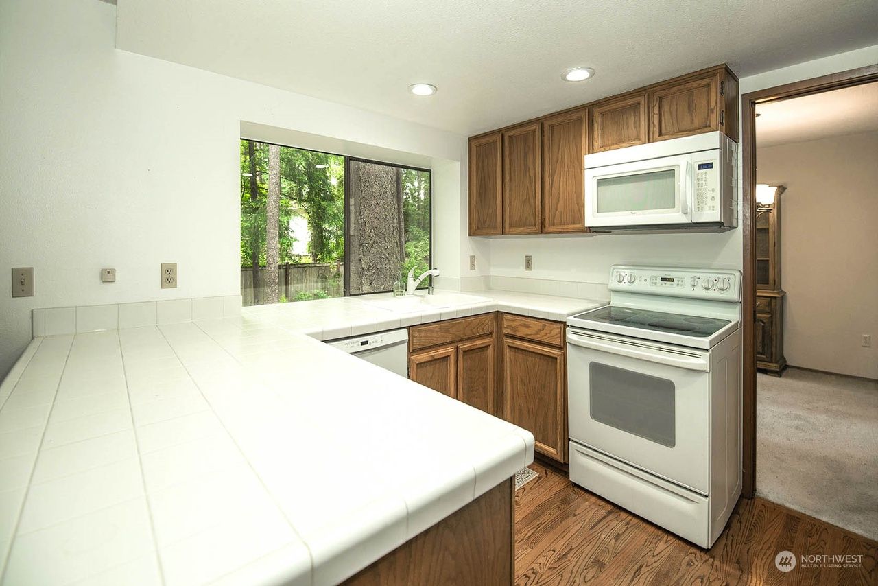 Bright kitchen with white counters, wood cabinets, white appliances, and a window over the sink