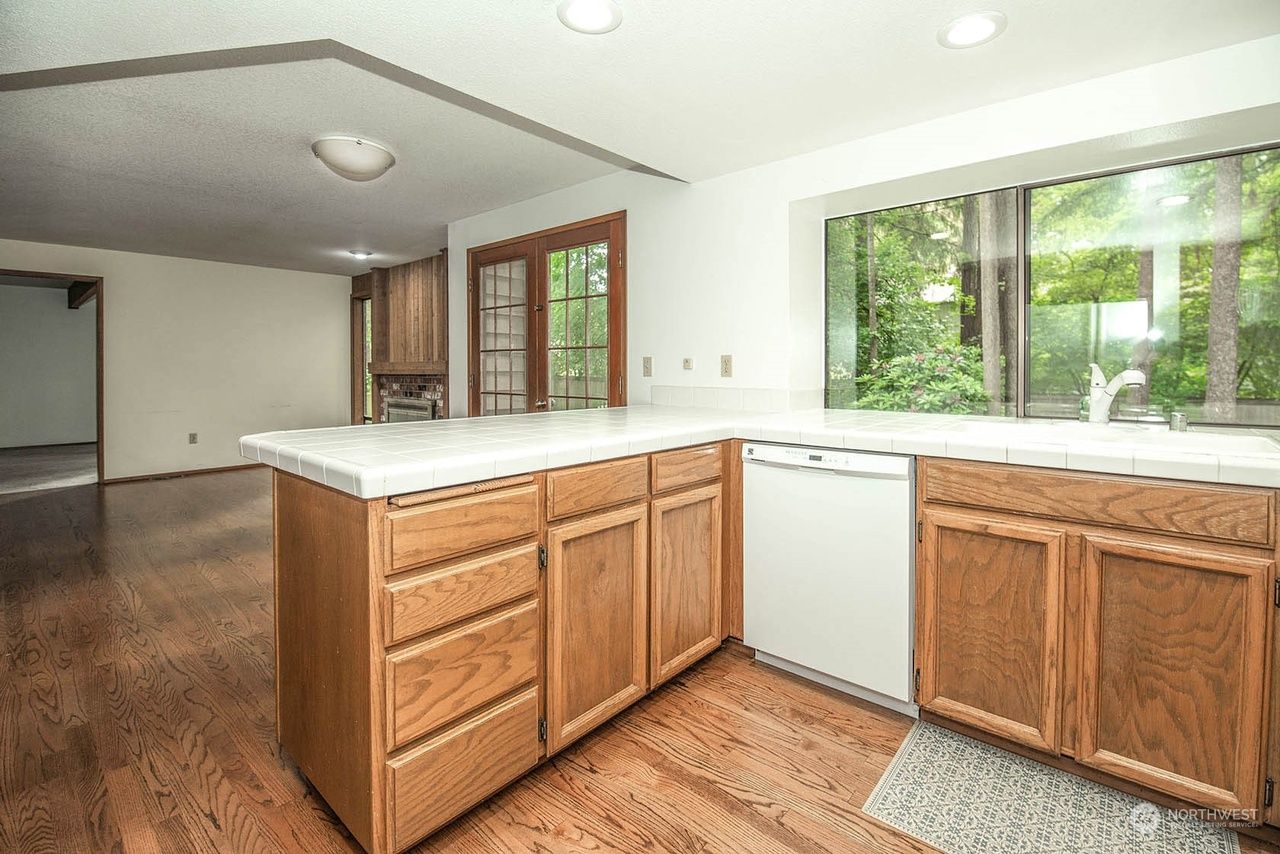 Empty kitchen with wood cabinets, white countertops, and a large window overlooking green trees.