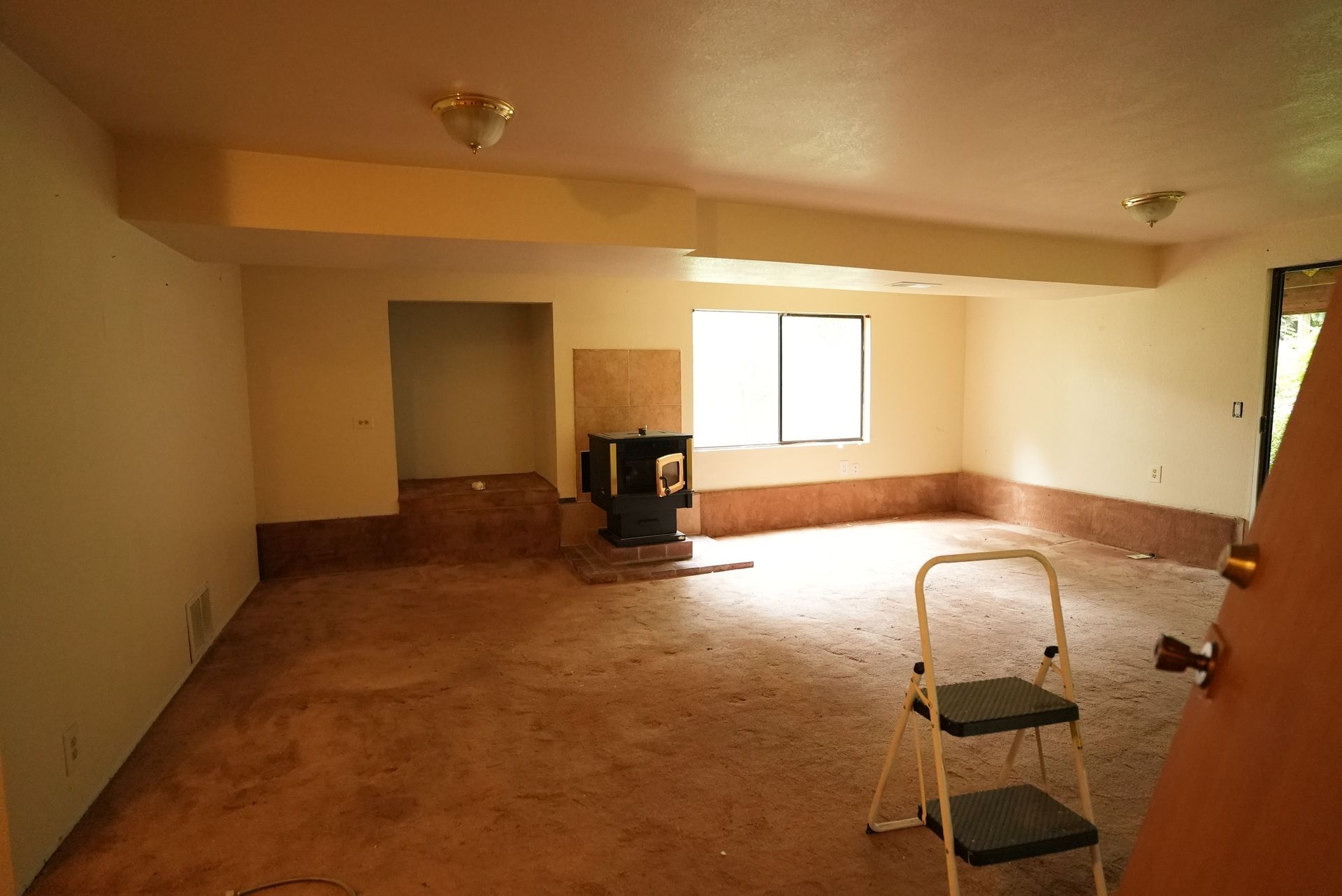 Empty beige living room with carpet, a window, and a step ladder near the foreground.