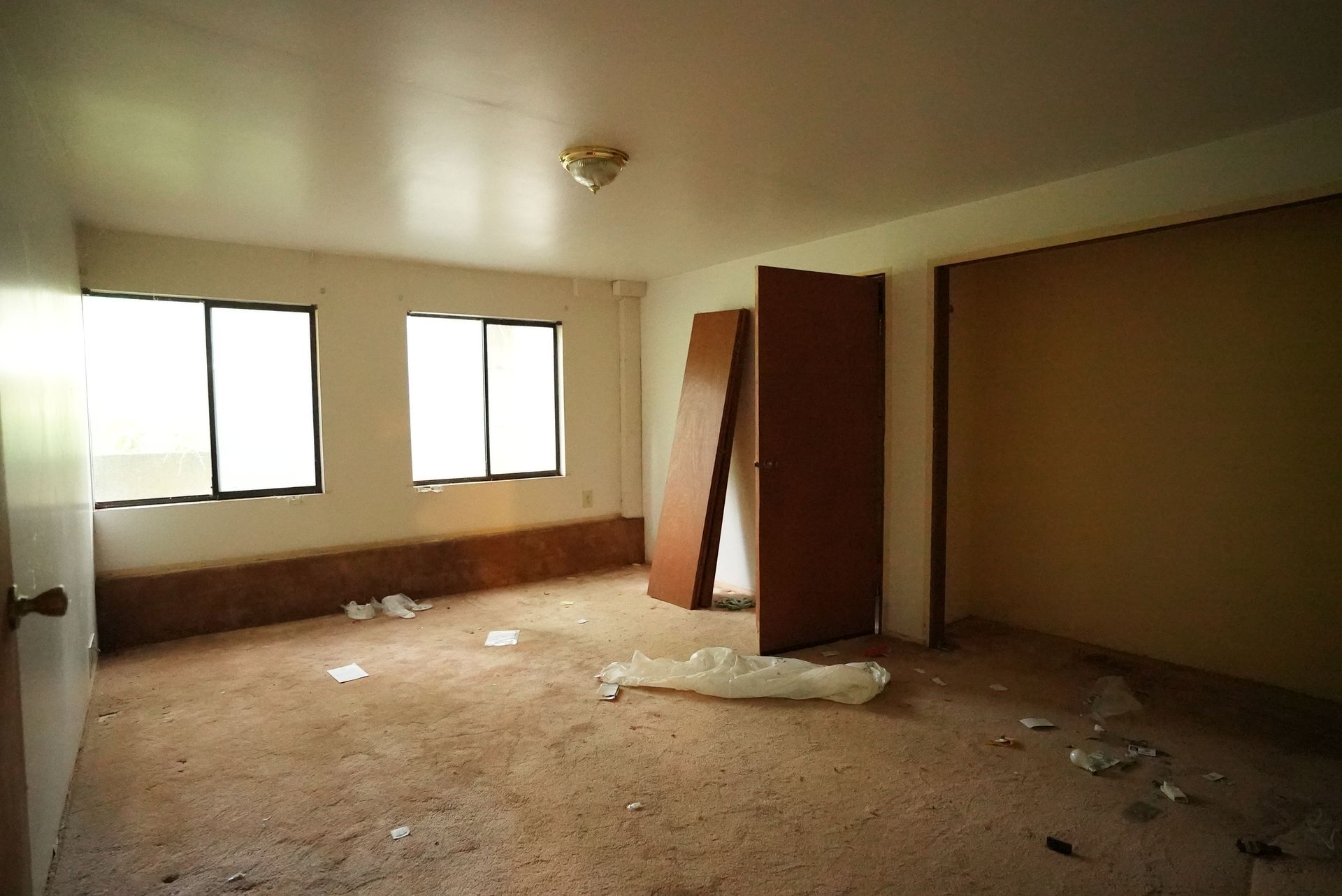 Empty, abandoned room with beige walls, windows, and debris scattered across the floor.