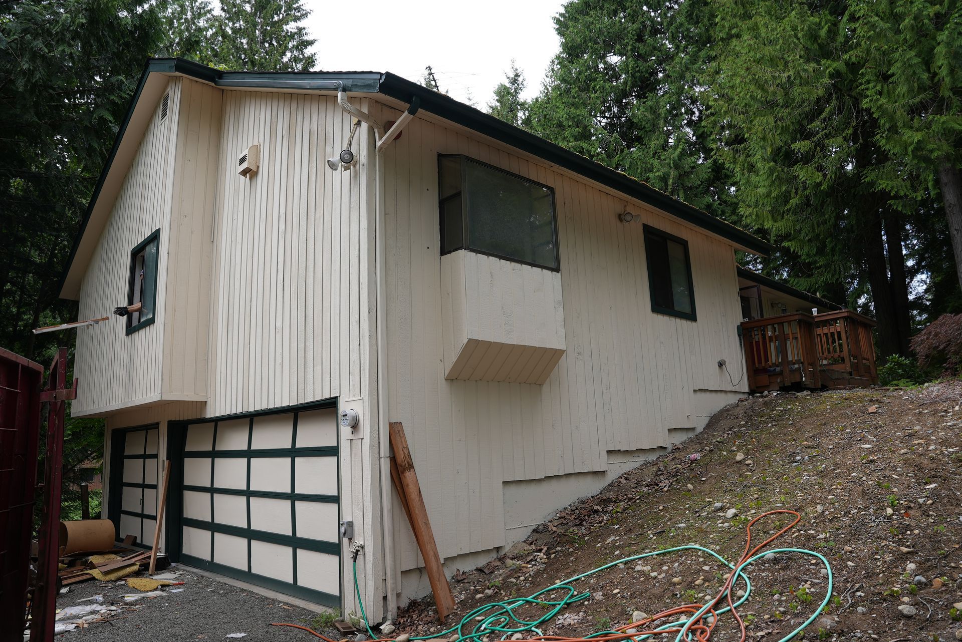 White house on a sloped lot with a garage and green-trimmed windows, surrounded by trees and gravel.