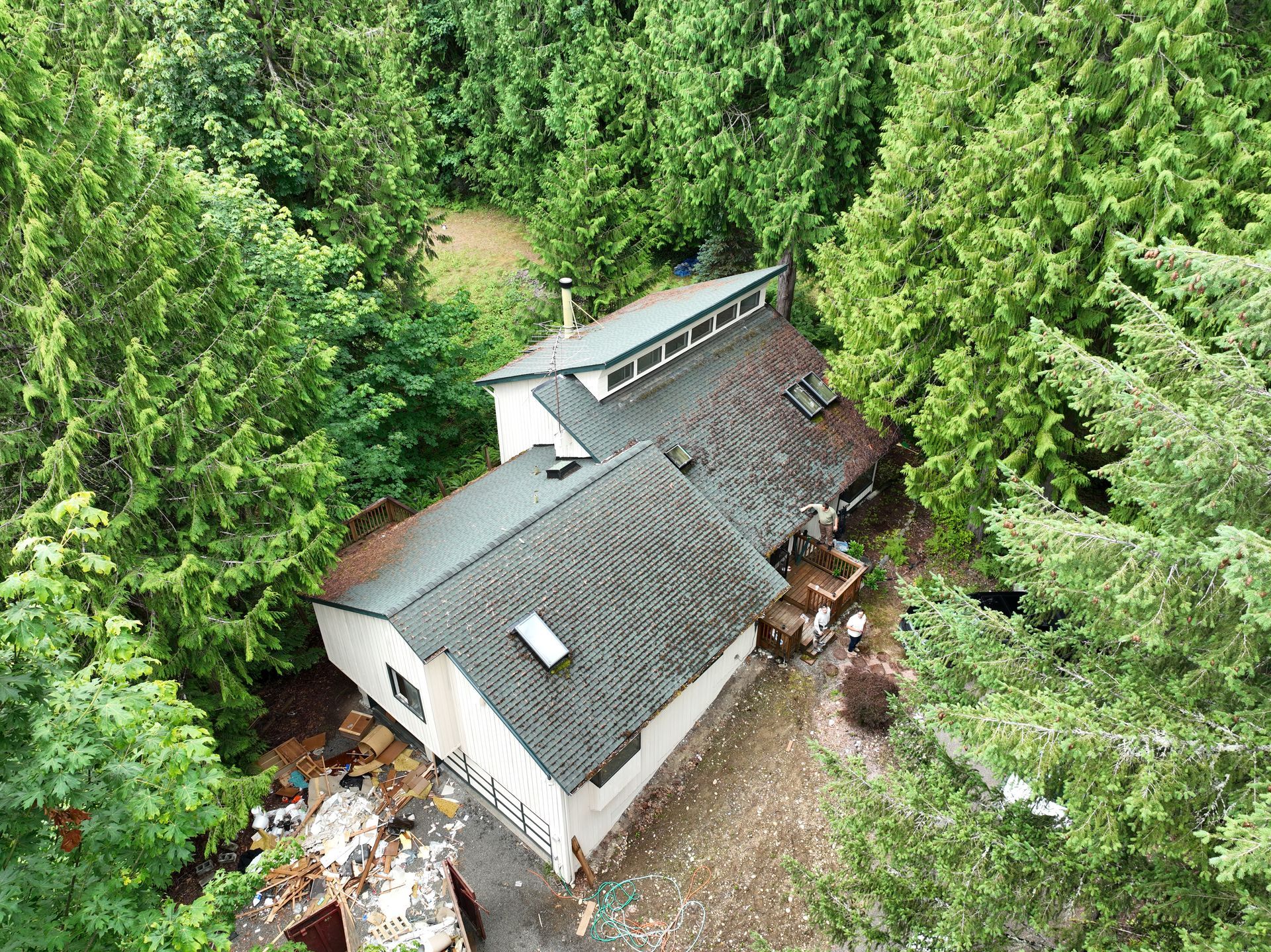 Aerial view of a small white cabin with a gray roof, surrounded by dense green forest.