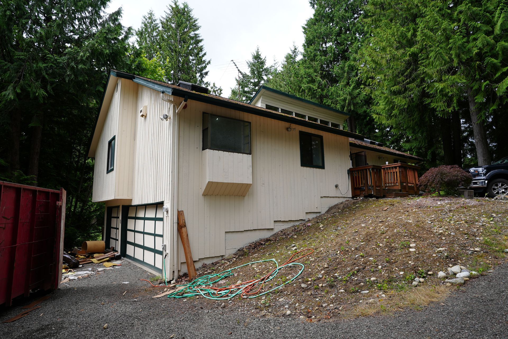 Small white house on a wooded hillside with a garage and gravel driveway