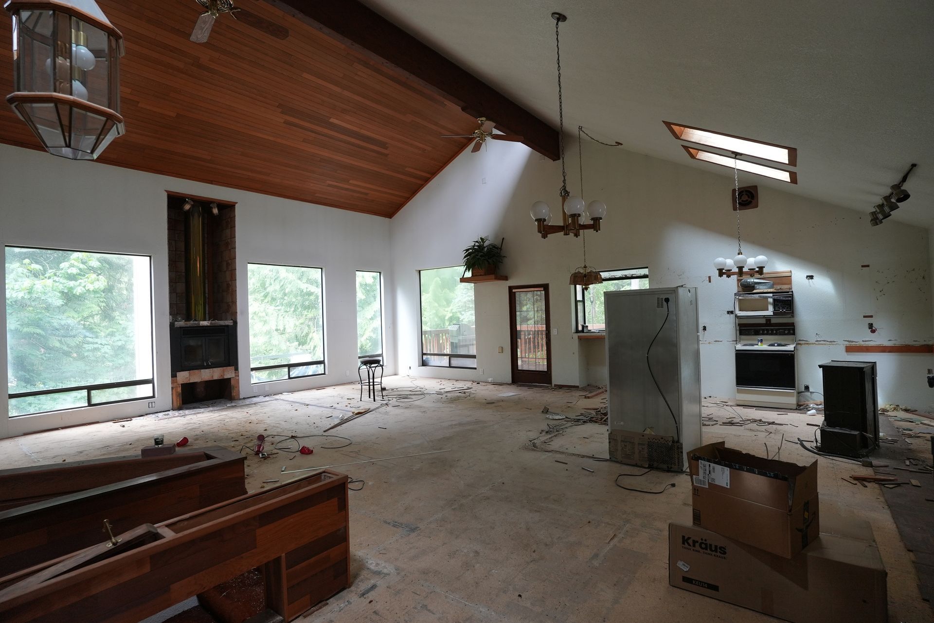 Abandoned living room with debris, vaulted wood ceiling, large windows, and a kitchen area.
