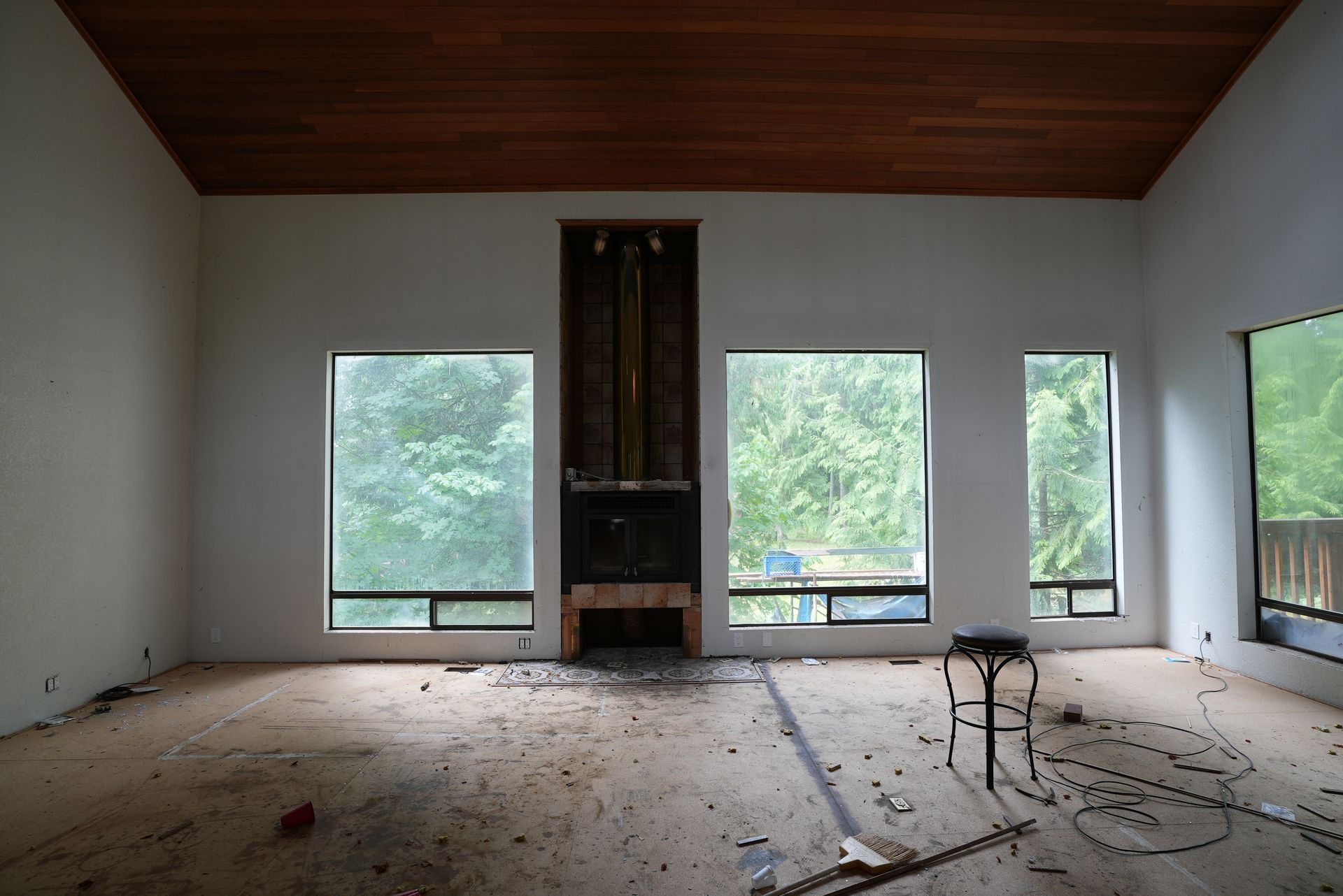 Abandoned living room with large windows, a central fireplace, and debris scattered across the floor