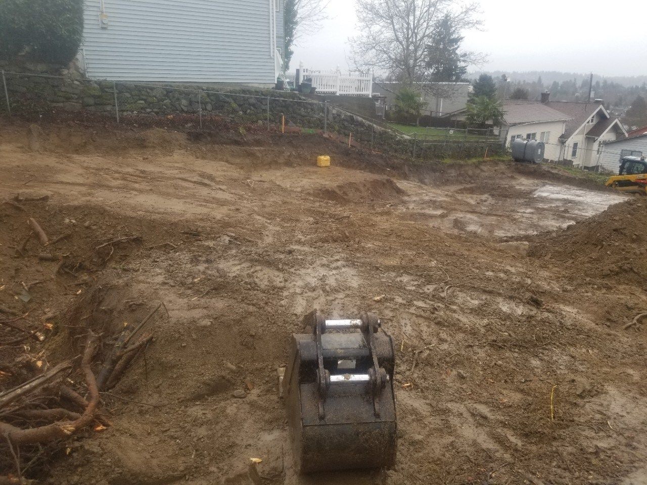 Construction site with a compact excavator on a muddy dirt lot near houses and trees.