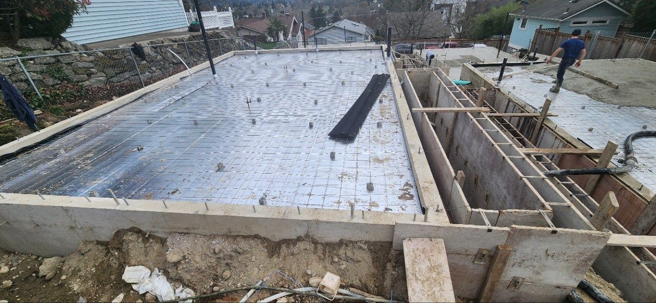 Construction site with a concrete foundation slab, rebar, wooden formwork, and workers in the background