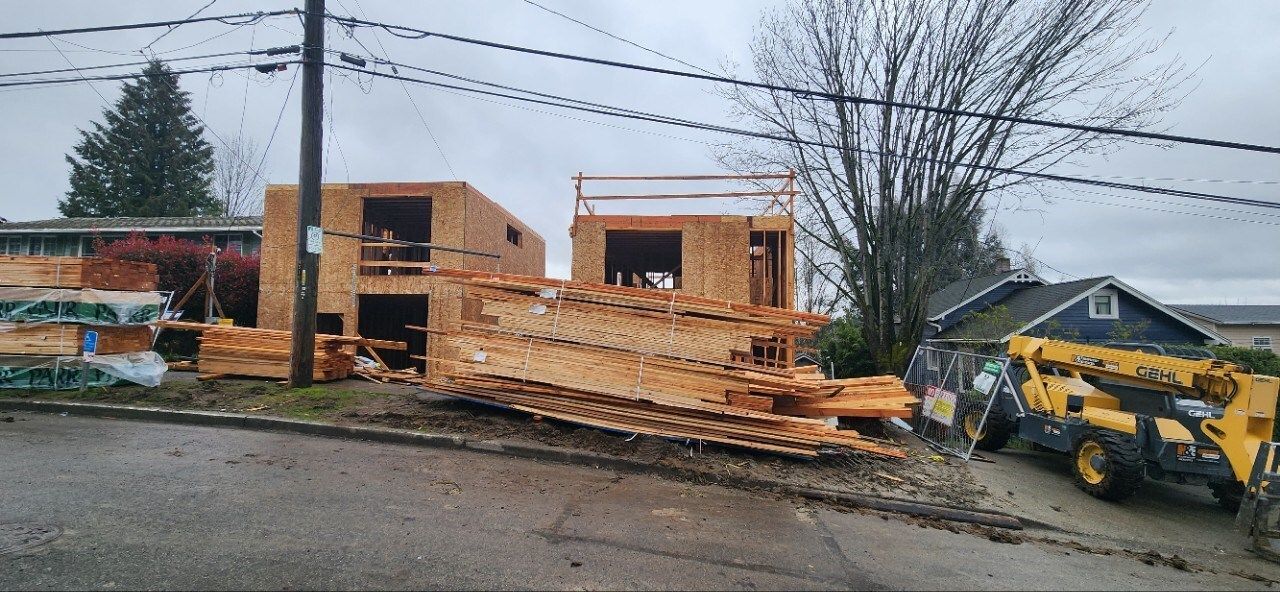 Pile of lumber at a construction site beside an excavator and unfinished brick building