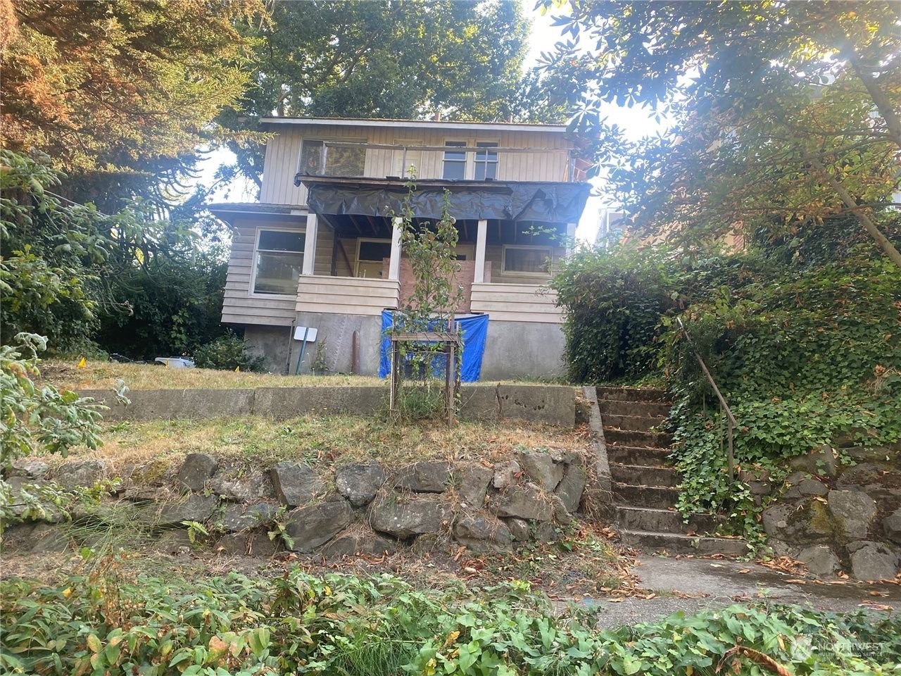 Abandoned two-story house behind a stone retaining wall, surrounded by dense trees and overgrown vegetation.