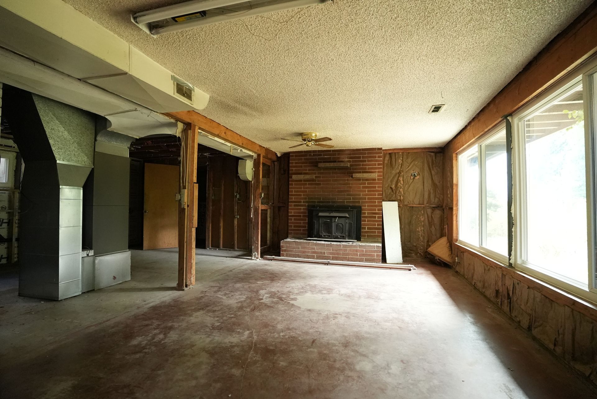 Empty living room with fireplace, large windows, exposed wood beams, and worn carpet.