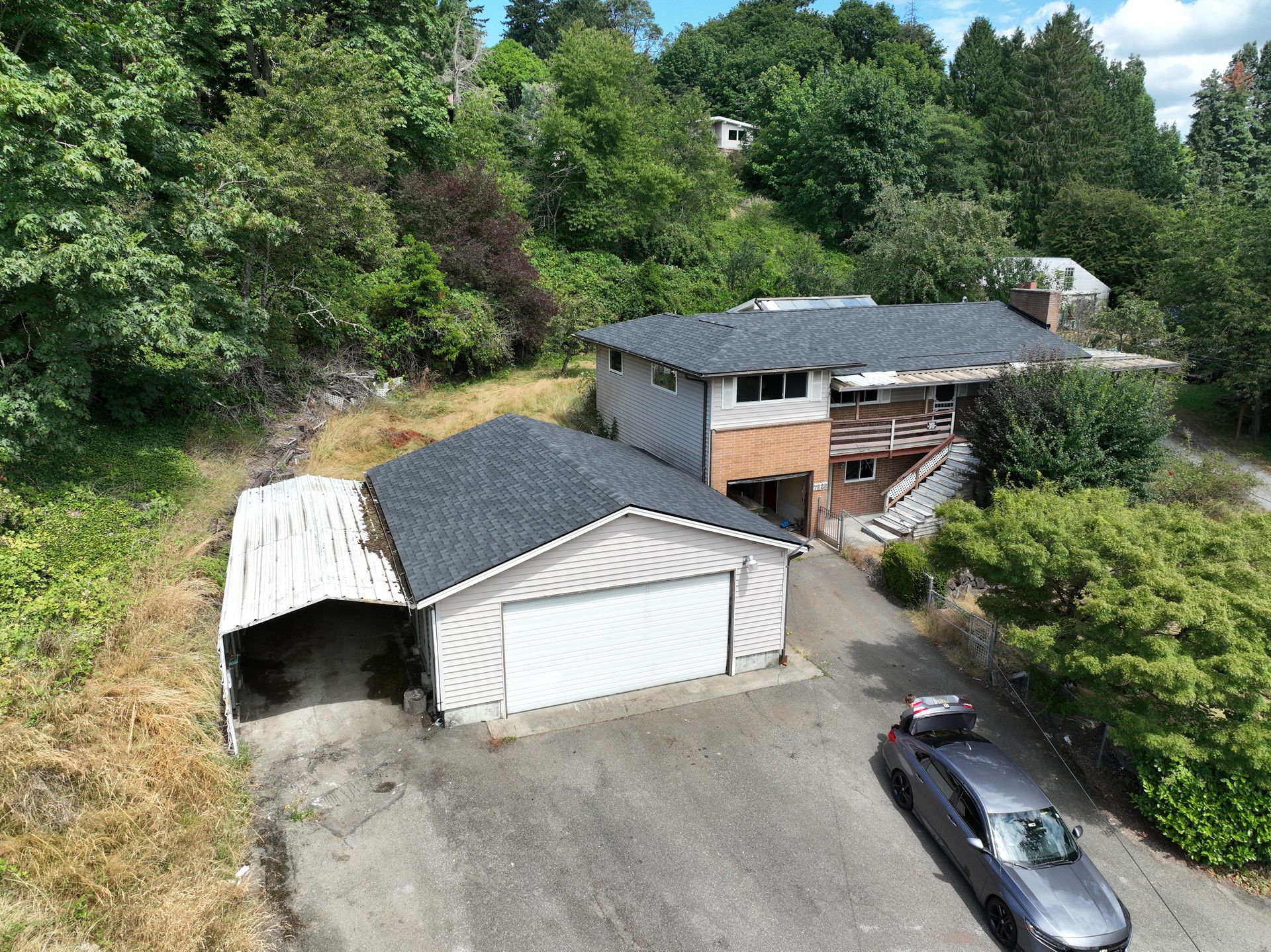 Aerial view of a house with detached garage, driveway, and parked car beside dense green trees.