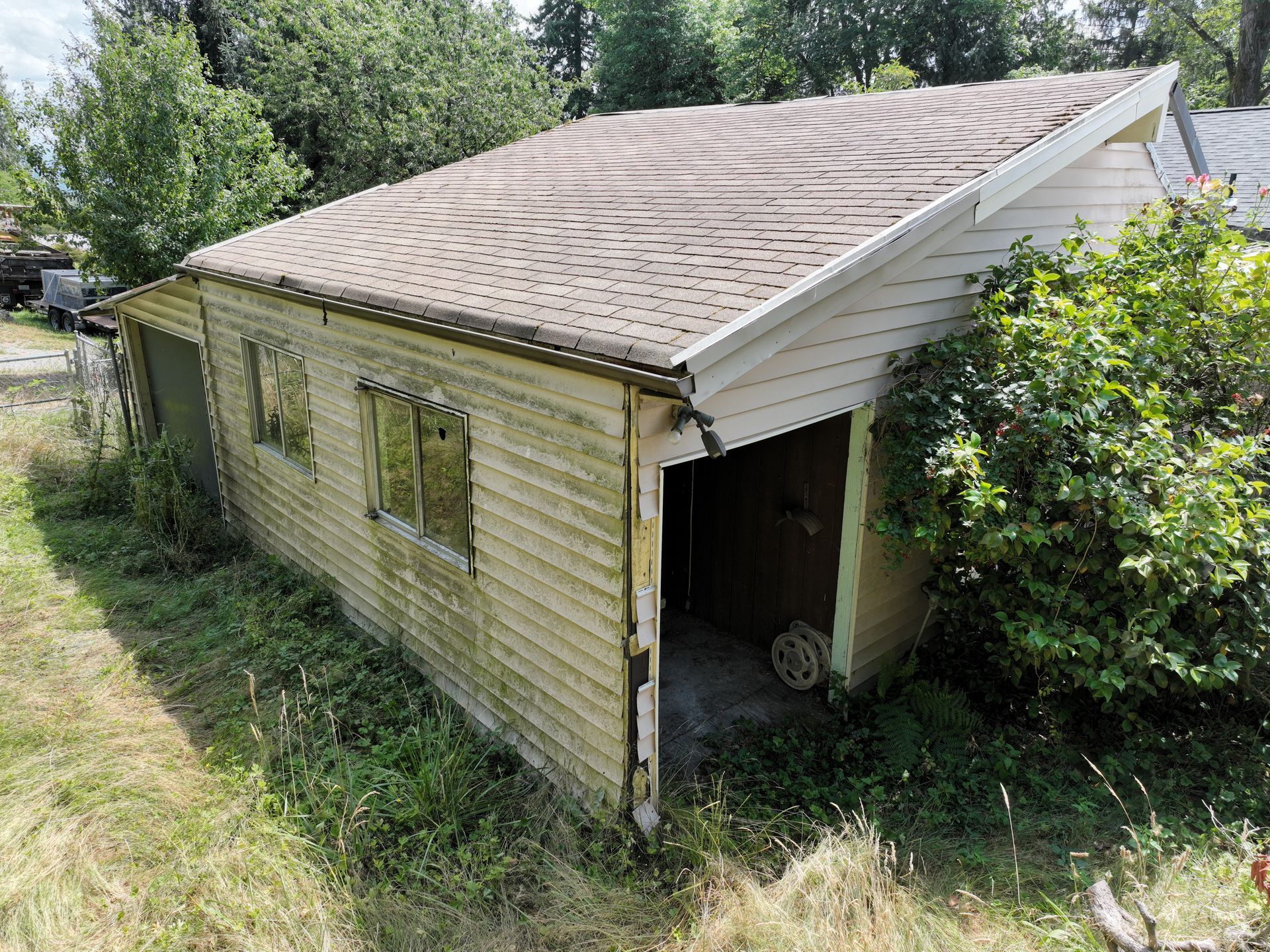 Abandoned beige house with a damaged roof, overgrown yard, and a dark open doorway