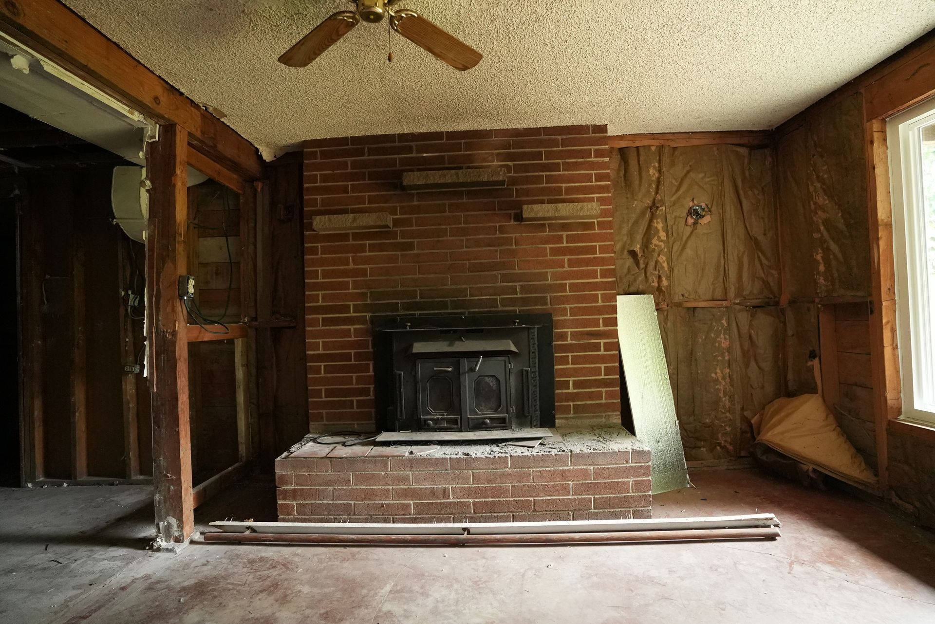 Empty living room with a brick fireplace, wood-paneled walls, and bare floorboards under a ceiling fan