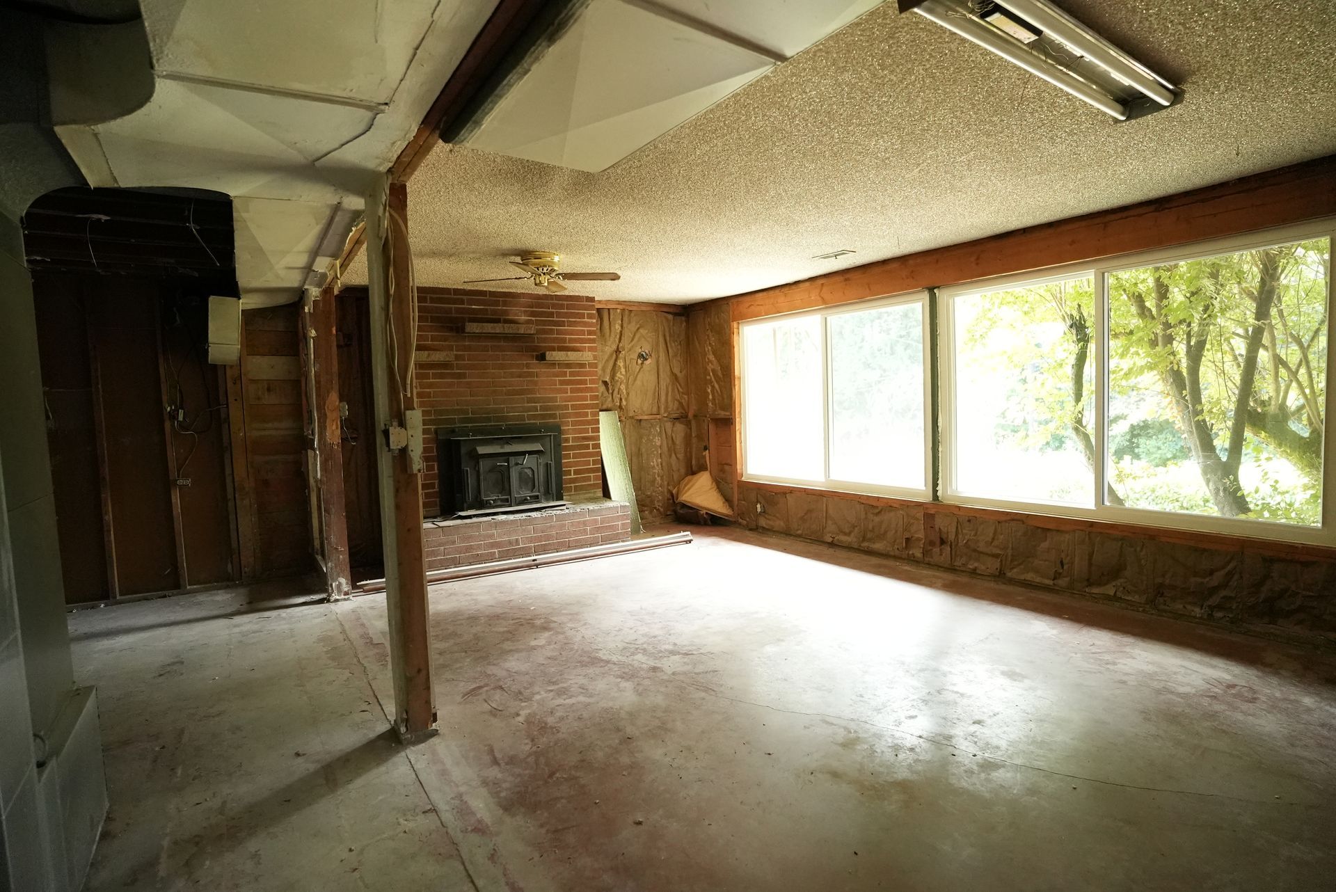 Empty sunlit living room with wood-paneled walls, fireplace, and large windows overlooking trees