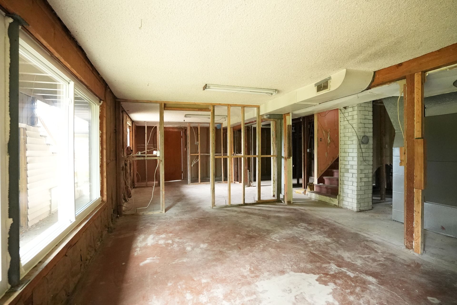 Empty room under renovation with exposed studs, worn floor, and large windows letting in daylight